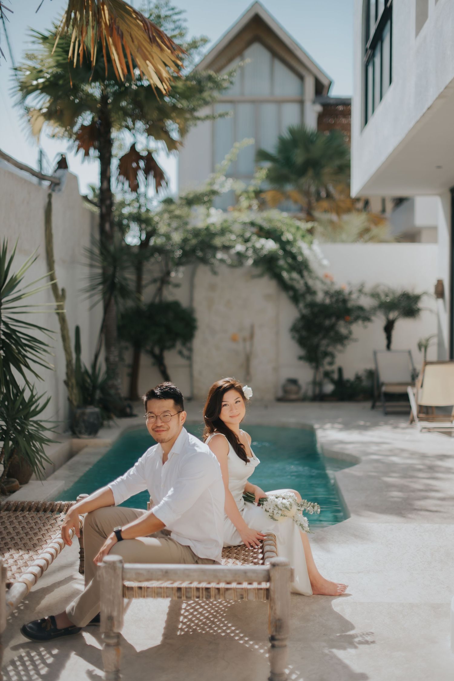Couple in white outfits sitting by a pool at a tropical villa, posing for an elegant pre-wedding photo shoot.