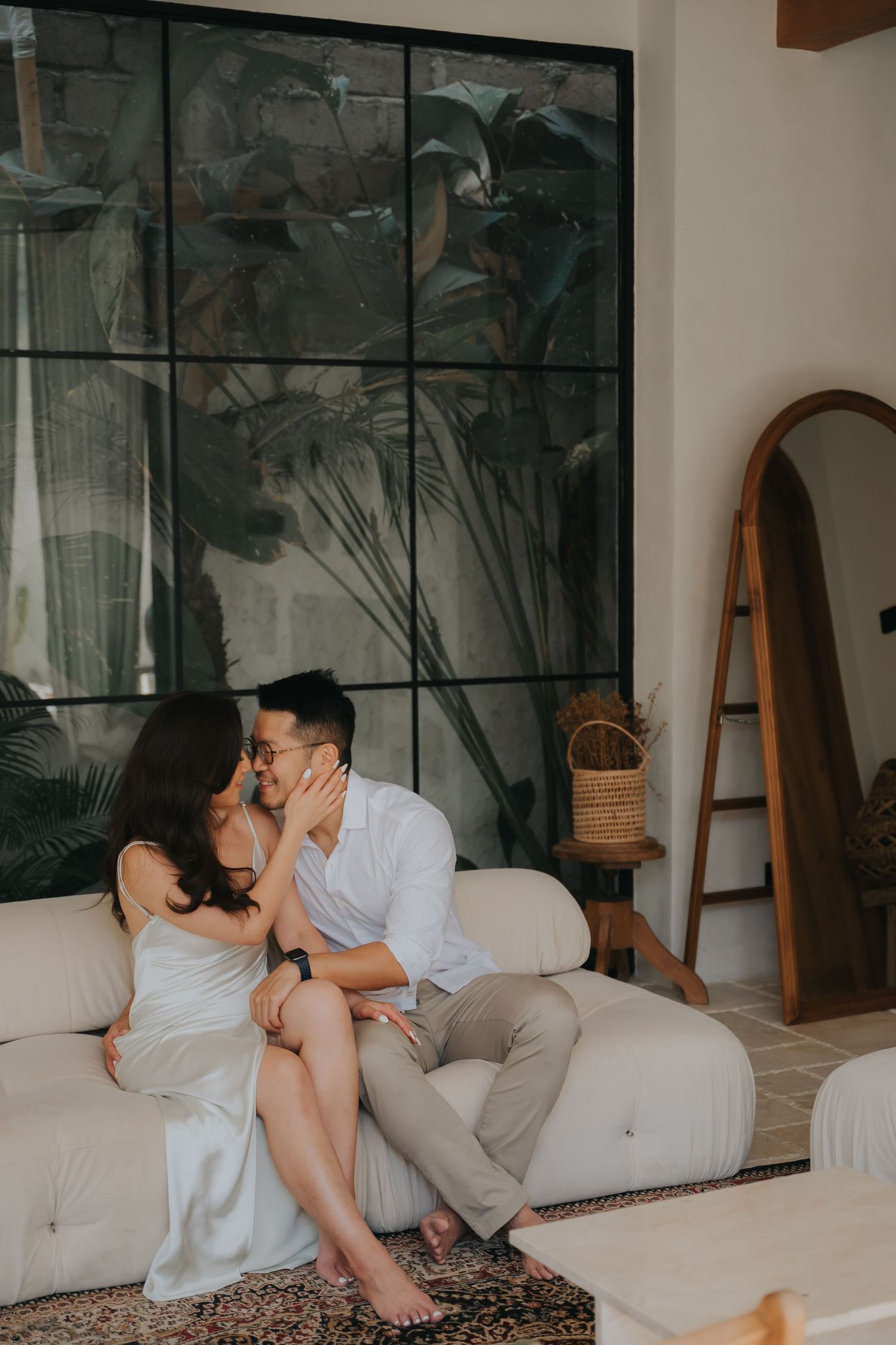 Couple sharing a romantic kiss on a white sofa in a modern boho living room with tropical plant wall art.