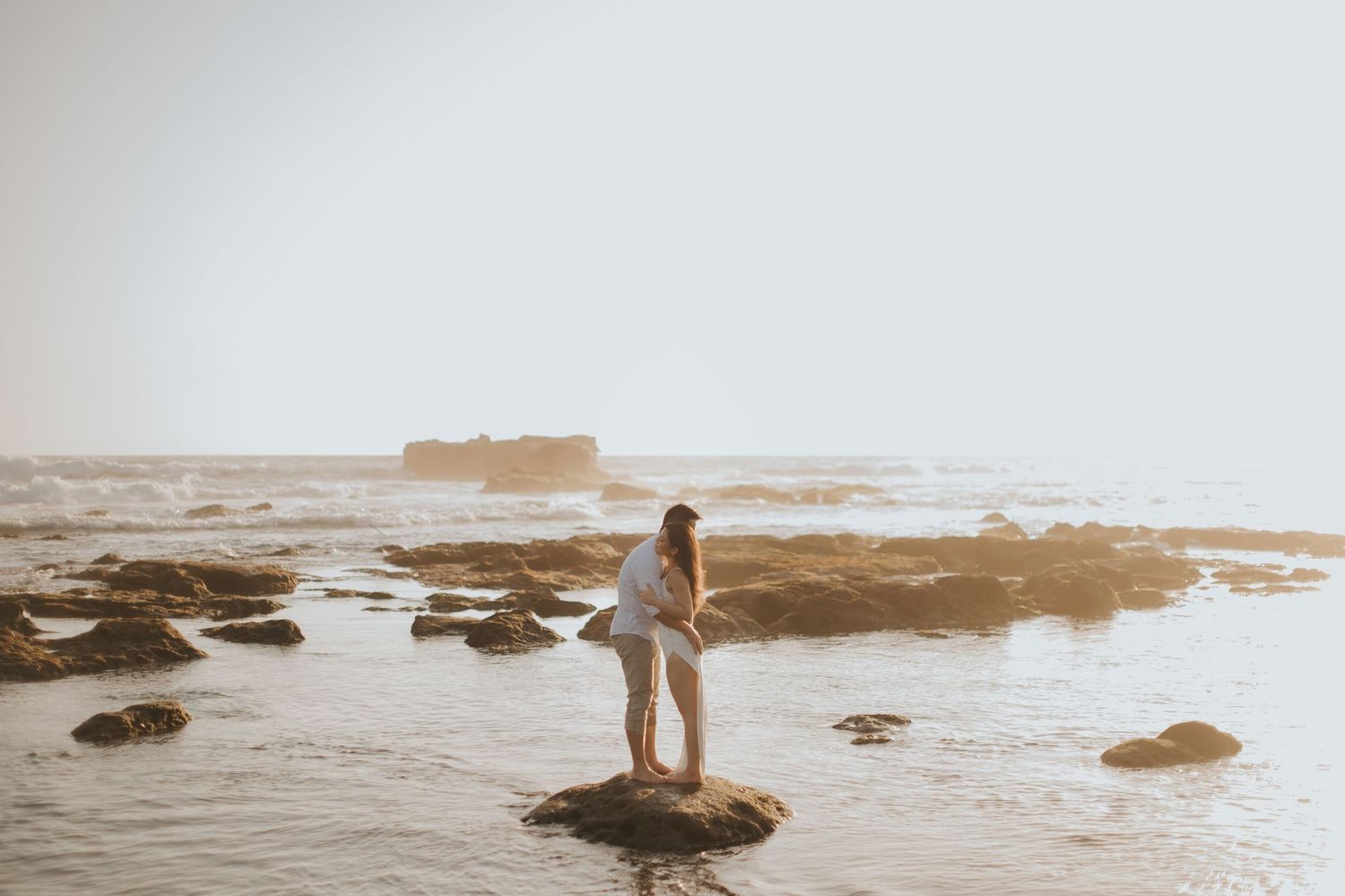 Couple embracing on rocky shore at sunset with calm ocean waves and misty horizon in background.