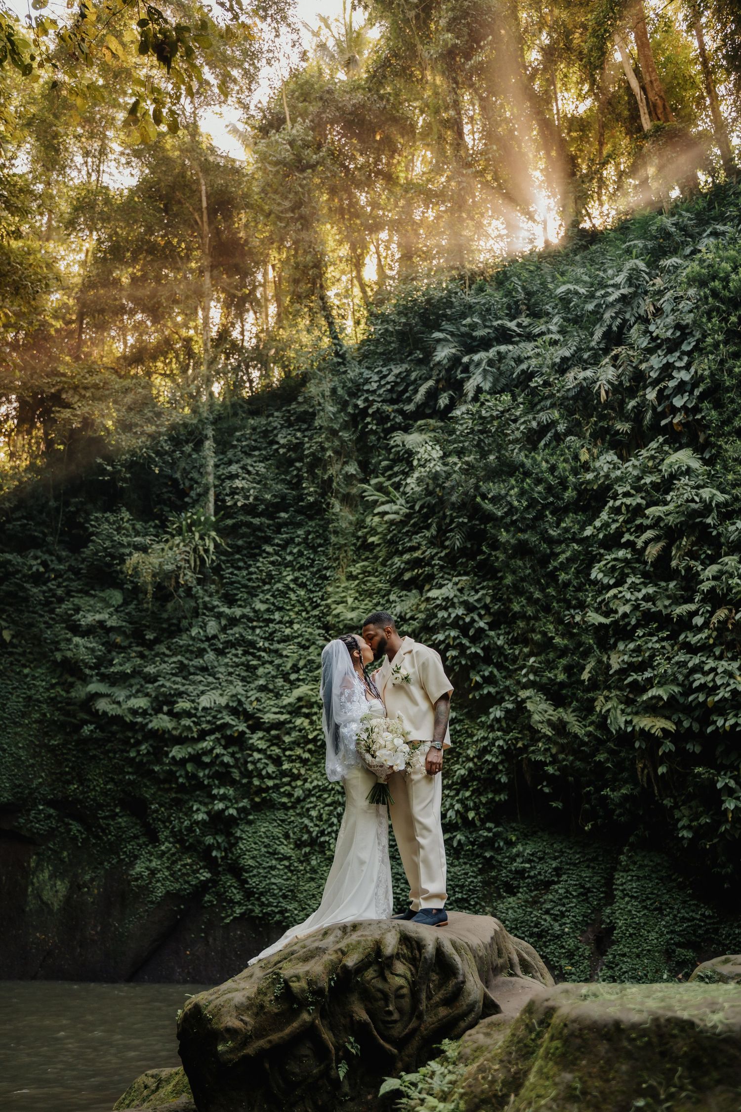 A bride and groom share a romantic kiss on a mossy rock surrounded by lush green forest with golden sunlight rays.