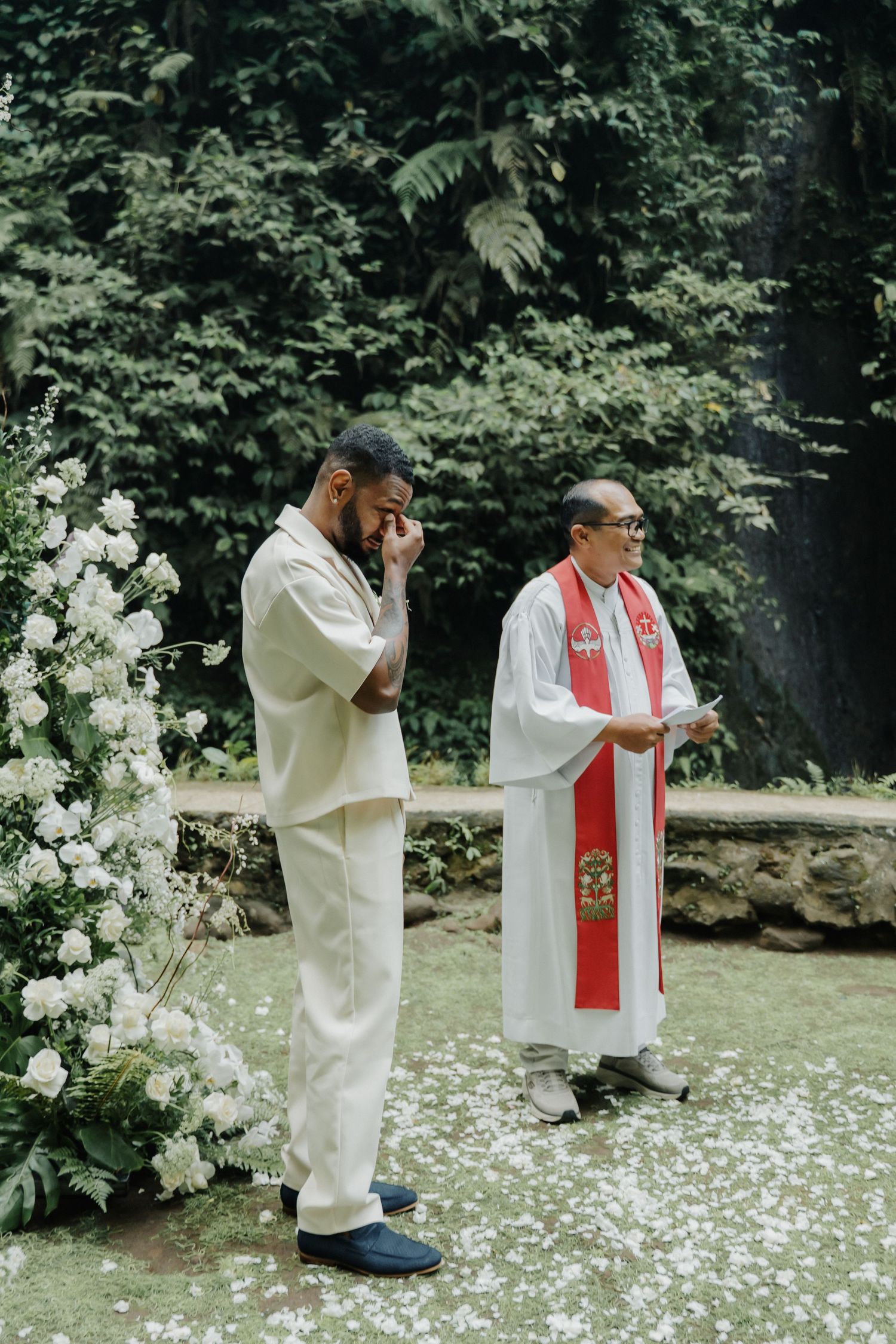 Emotional groom wiping tears beside officiant during an outdoor wedding ceremony with white floral arch.