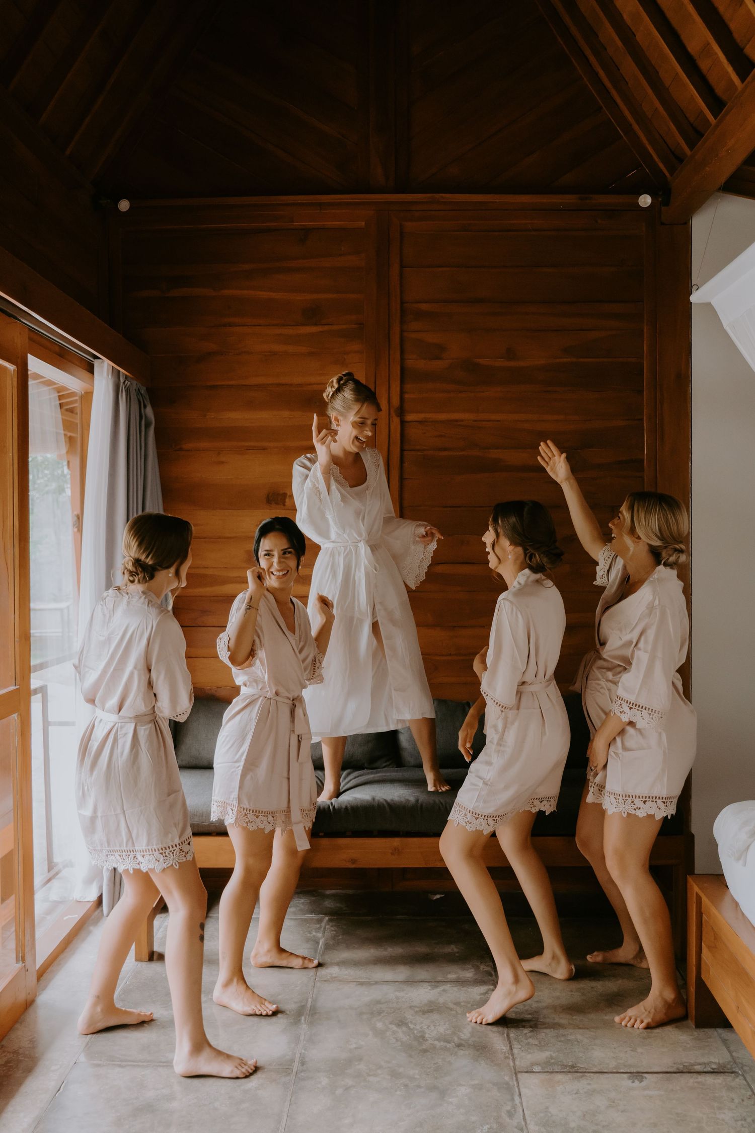 Bridesmaids in matching blush robes celebrate with bride jumping on couch in wooden Bali villa.