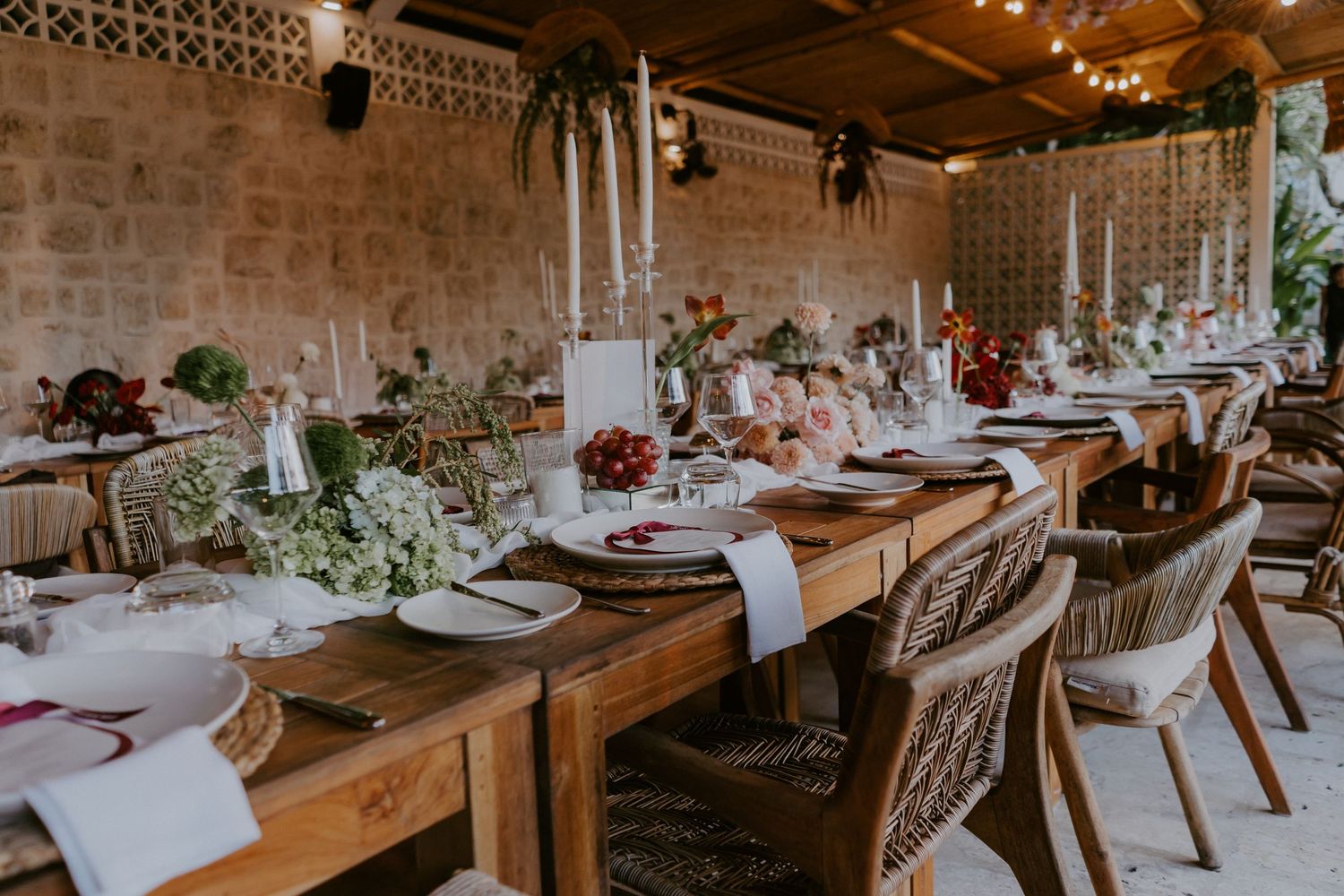 Rustic wedding reception table with tall white candles, floral centerpieces, and wicker chairs in stone venue.