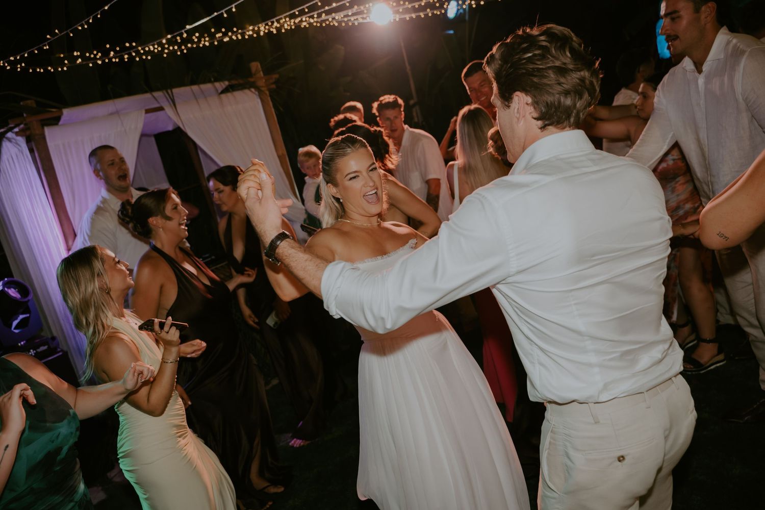 Bride and groom dance joyfully with guests at lively outdoor wedding reception at night.