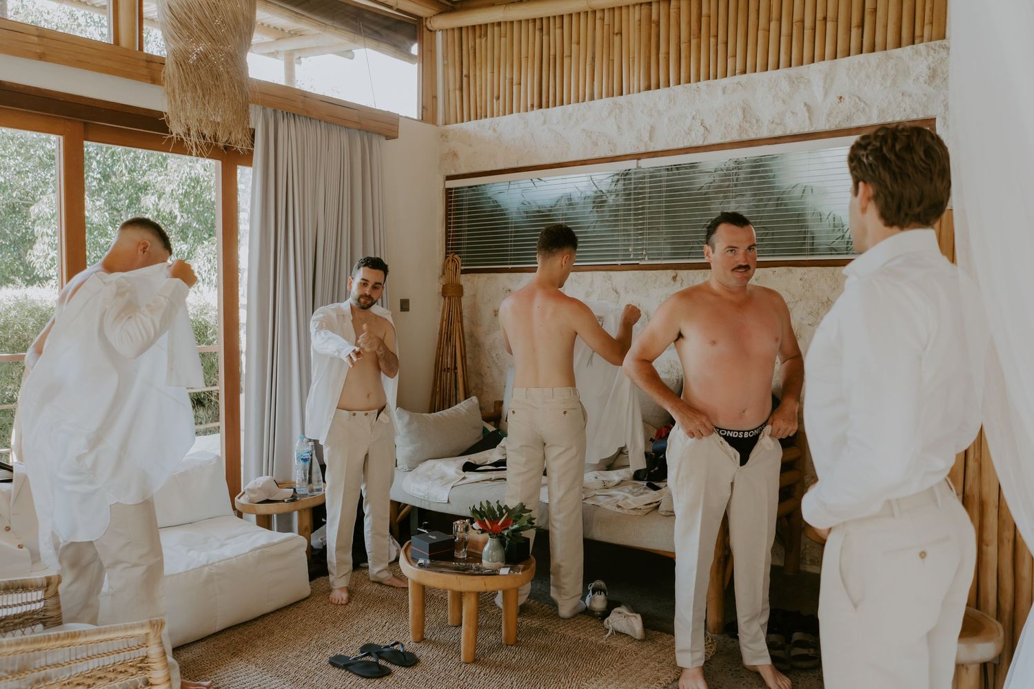 Groomsmen getting ready in a bright villa room, some shirtless in white pants before a wedding ceremony.