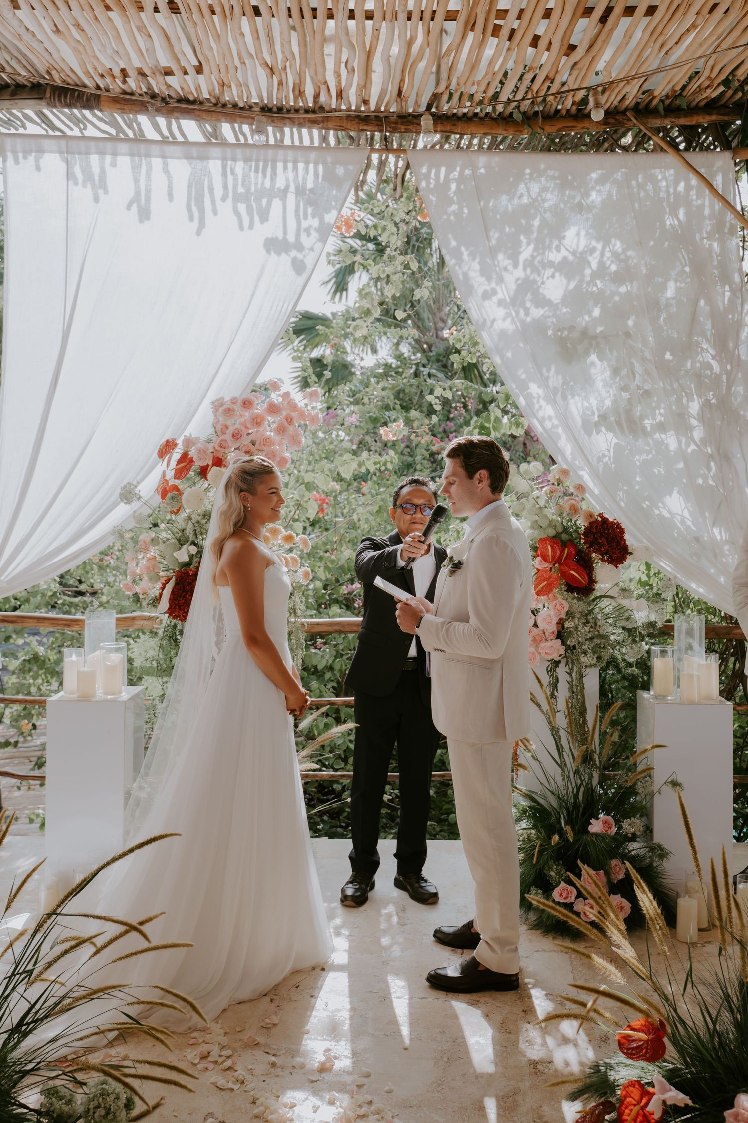 Bride and groom exchange vows under a floral arch with white draping and tropical garden backdrop.