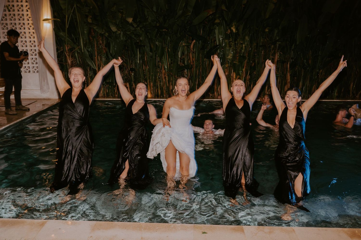 Bride celebrating in pool with bridesmaids in black dresses raising arms joyfully at night.