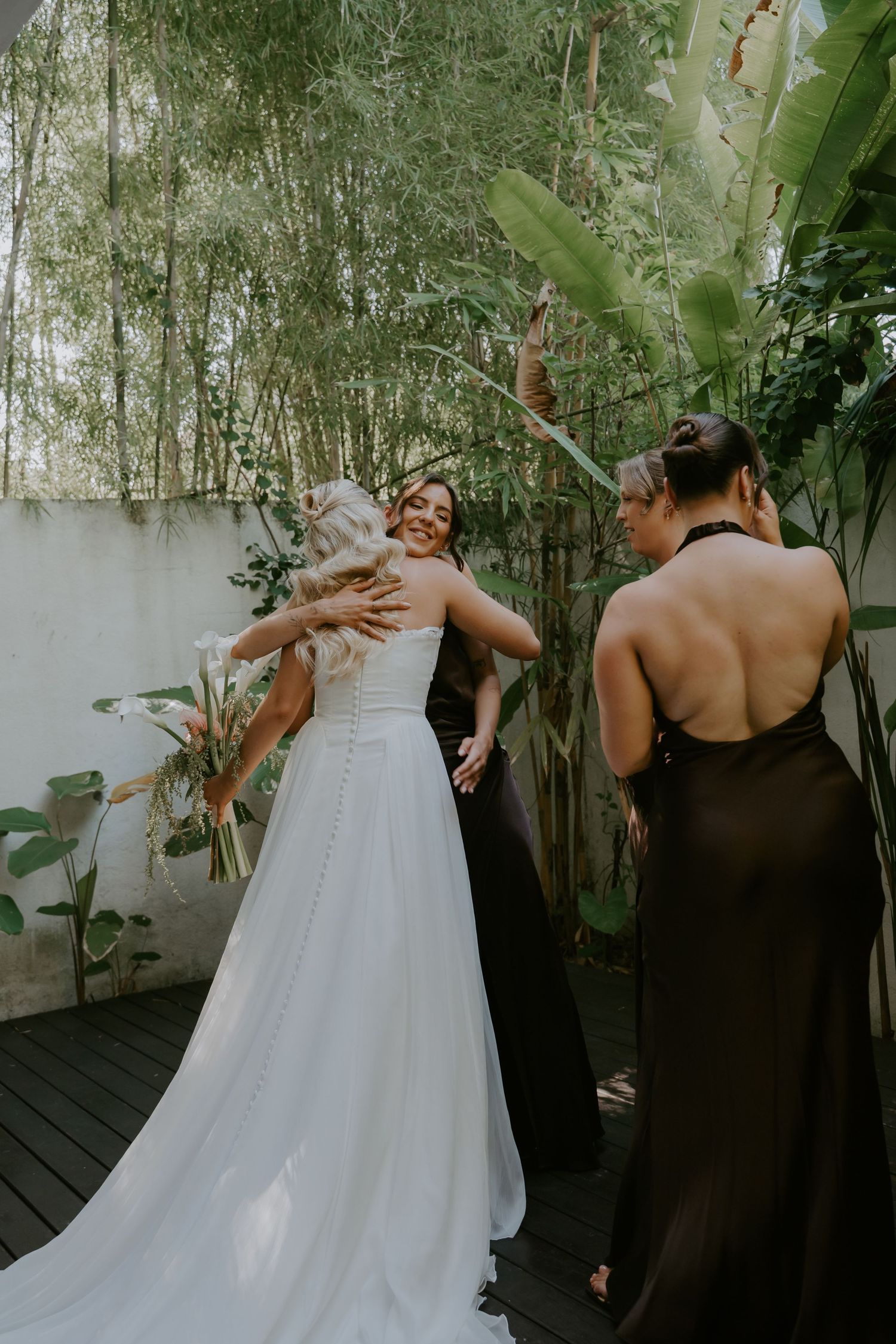 Bride in elegant white A-line wedding dress being assisted by bridesmaids in black halter gowns outdoors.