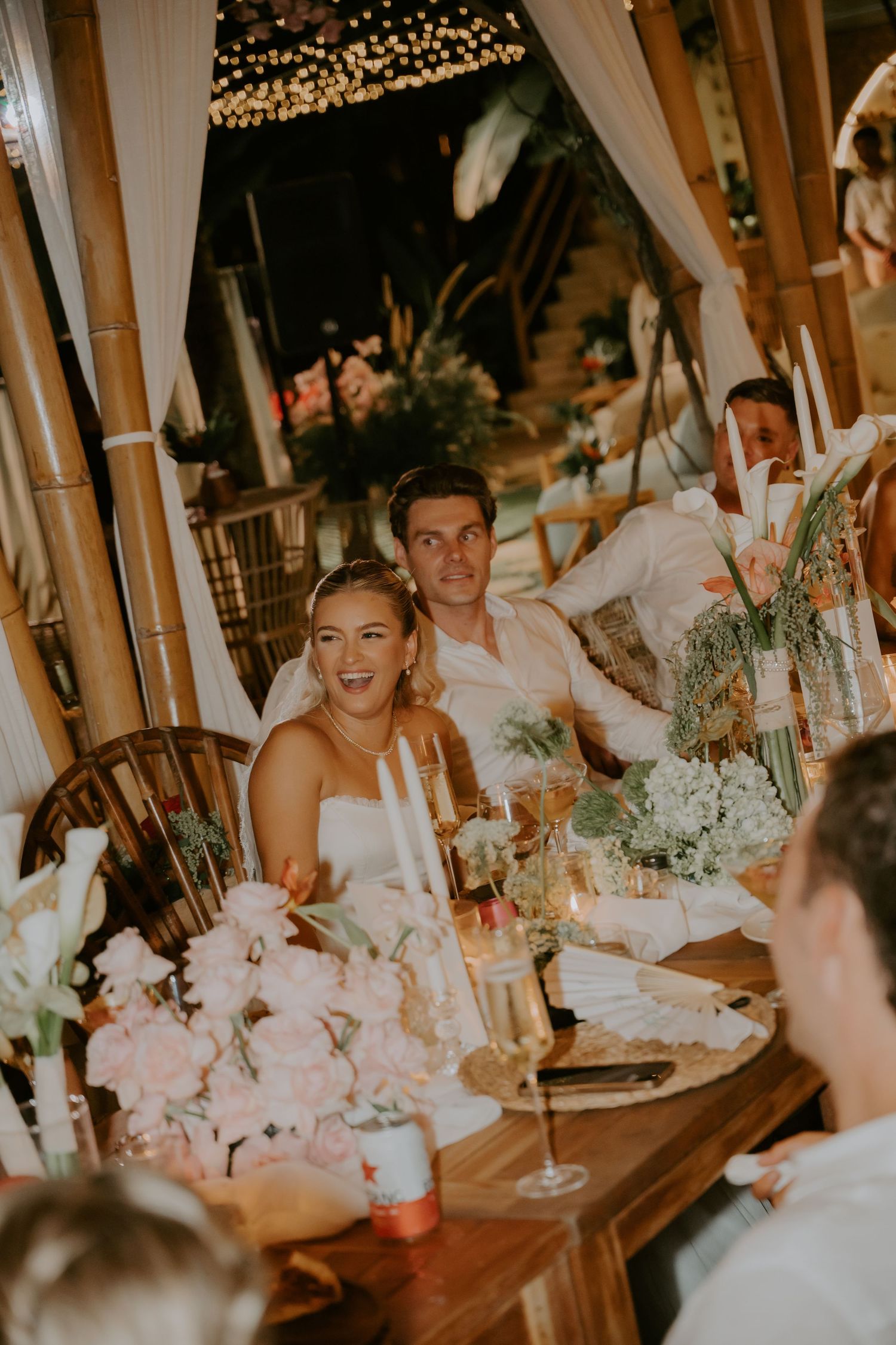 Smiling bride and groom seated at a floral decorated wedding reception table with candlelight.