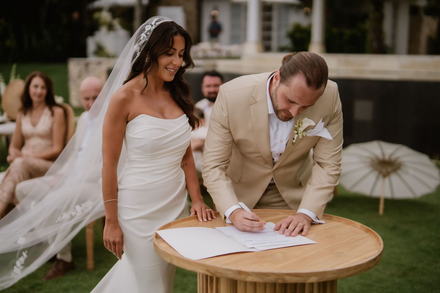 Bride smiling as groom signs wedding certificate at wooden table during outdoor garden ceremony.