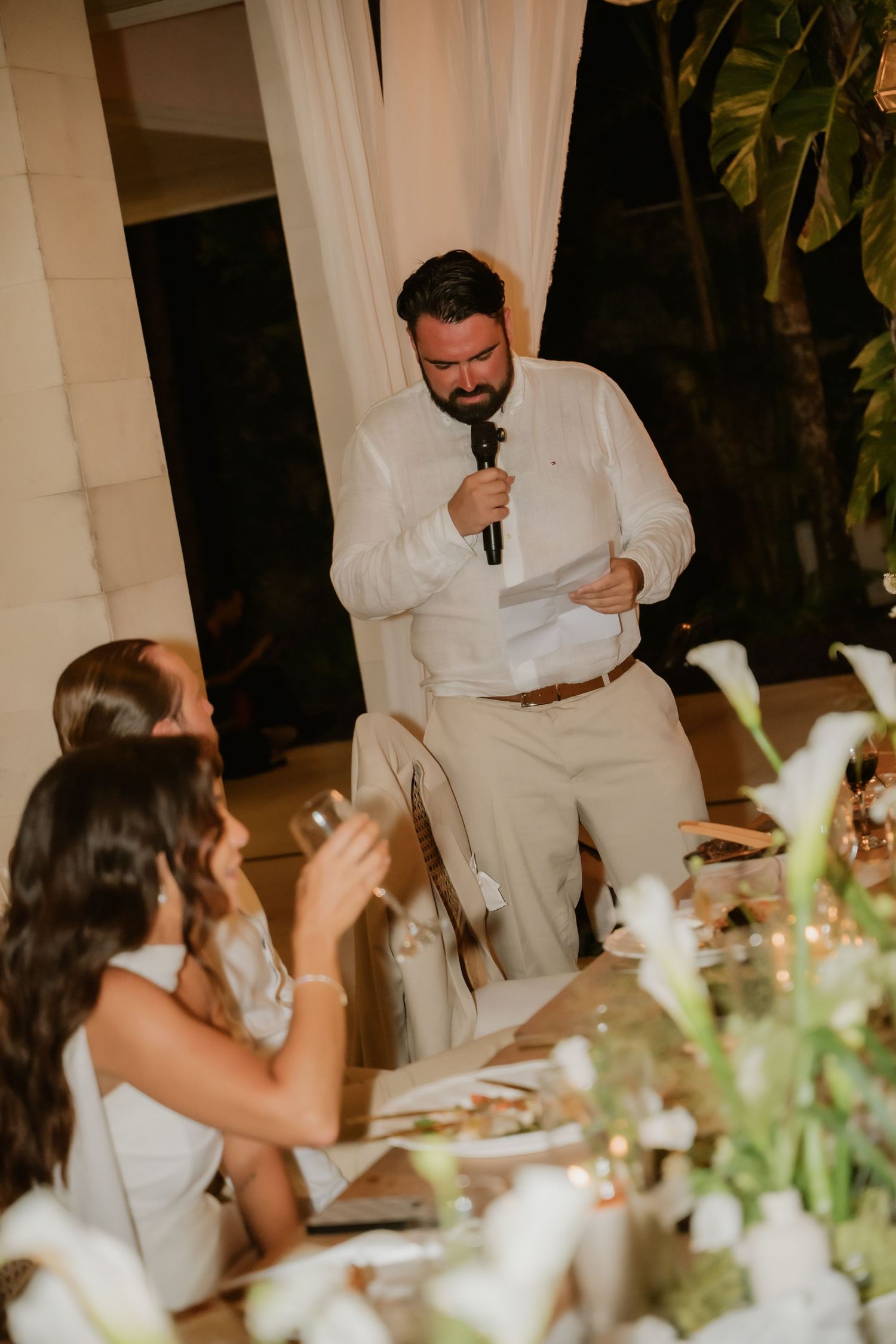 Man in white shirt delivers heartfelt speech with microphone at outdoor wedding reception dinner.
