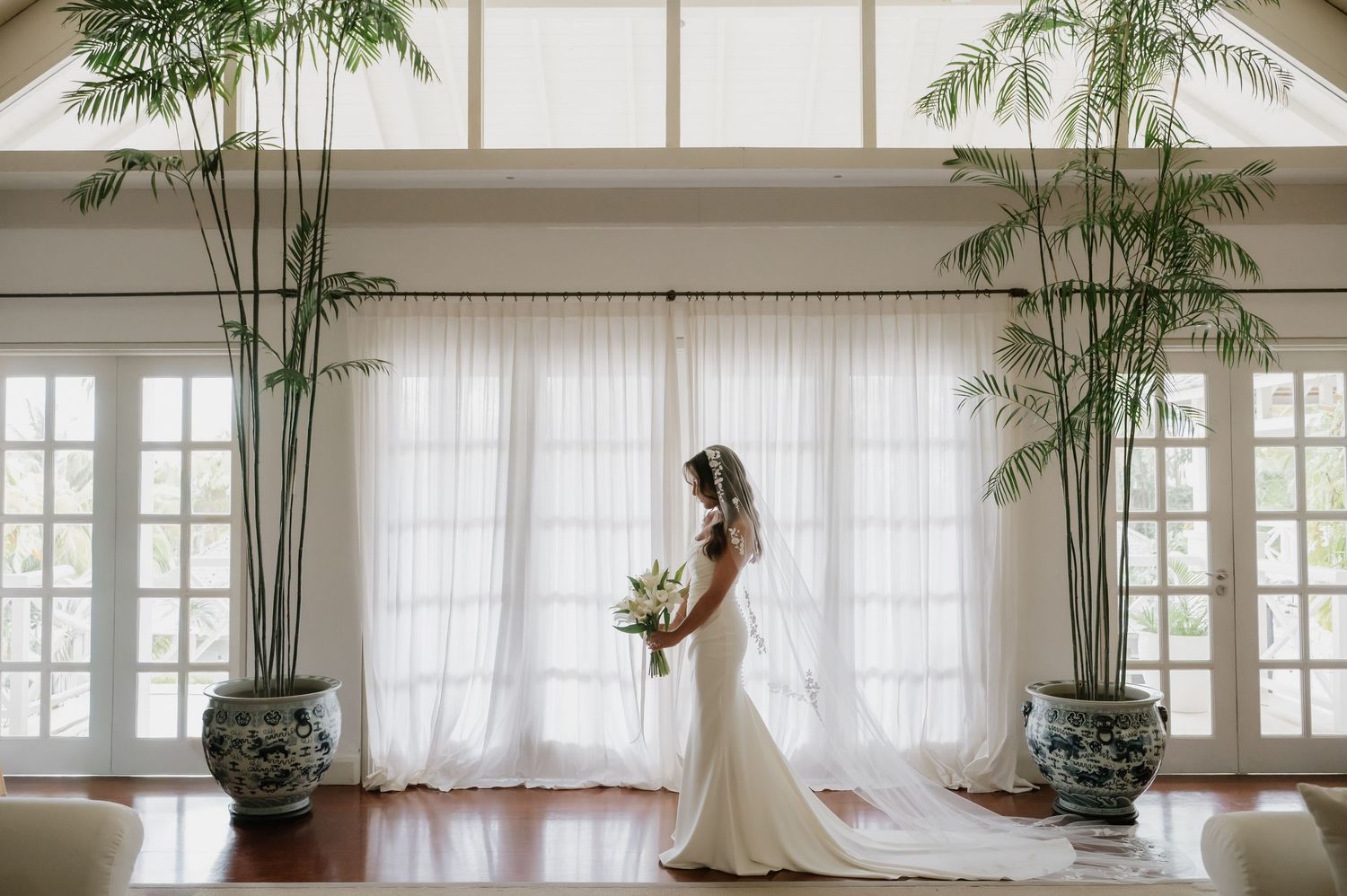 Bride in white gown holding bouquet stands in elegant room with tall palms and sheer curtains.