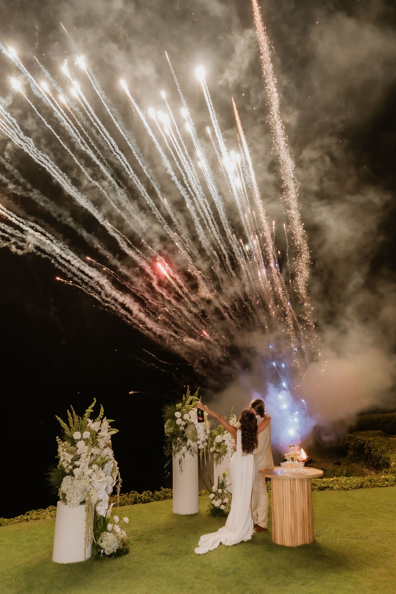 Bride and groom share kiss as spectacular fireworks explode over cliffside wedding ceremony at night.