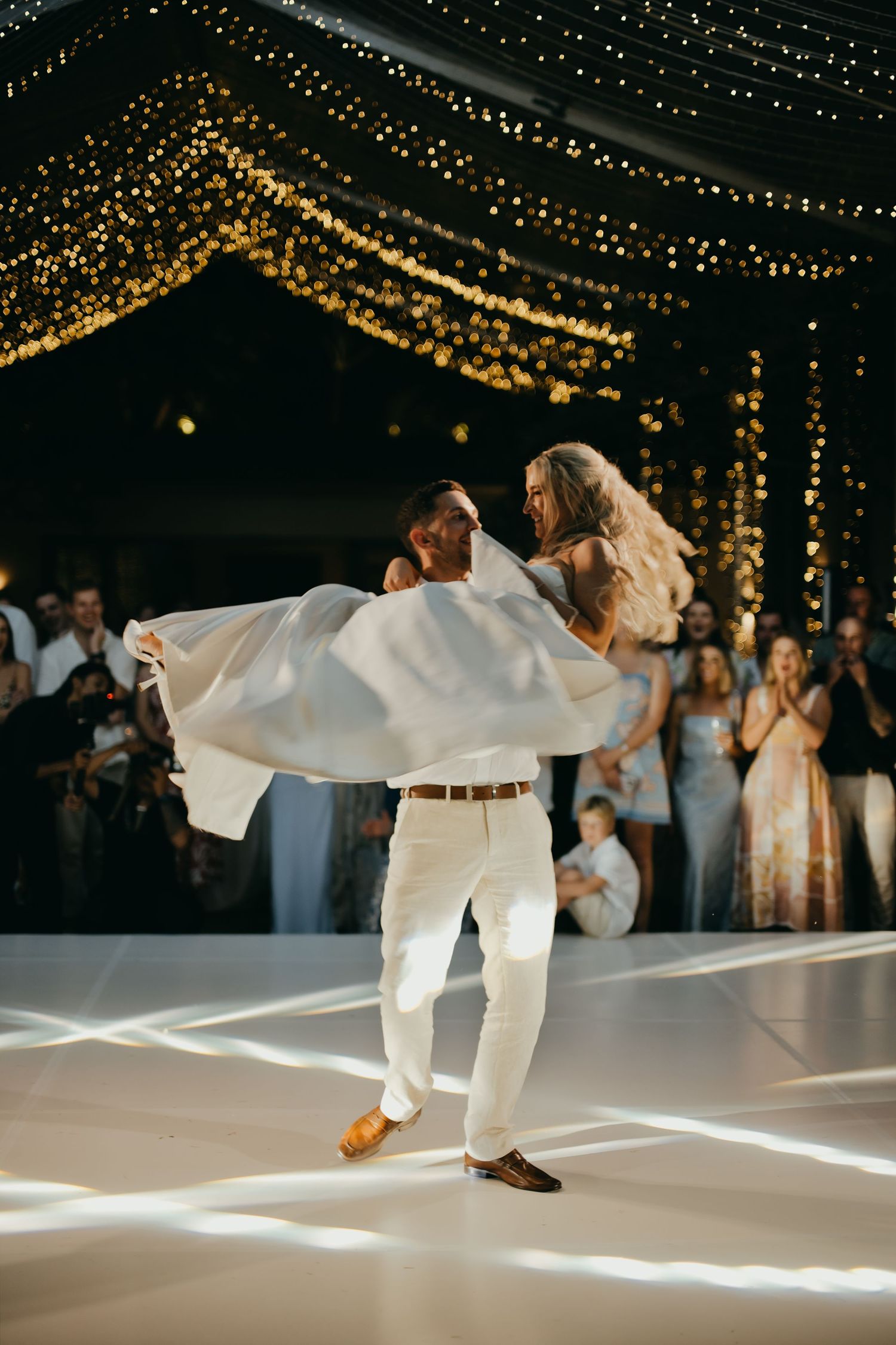 Groom lifts bride in white dress during first dance under golden fairy lights at reception.