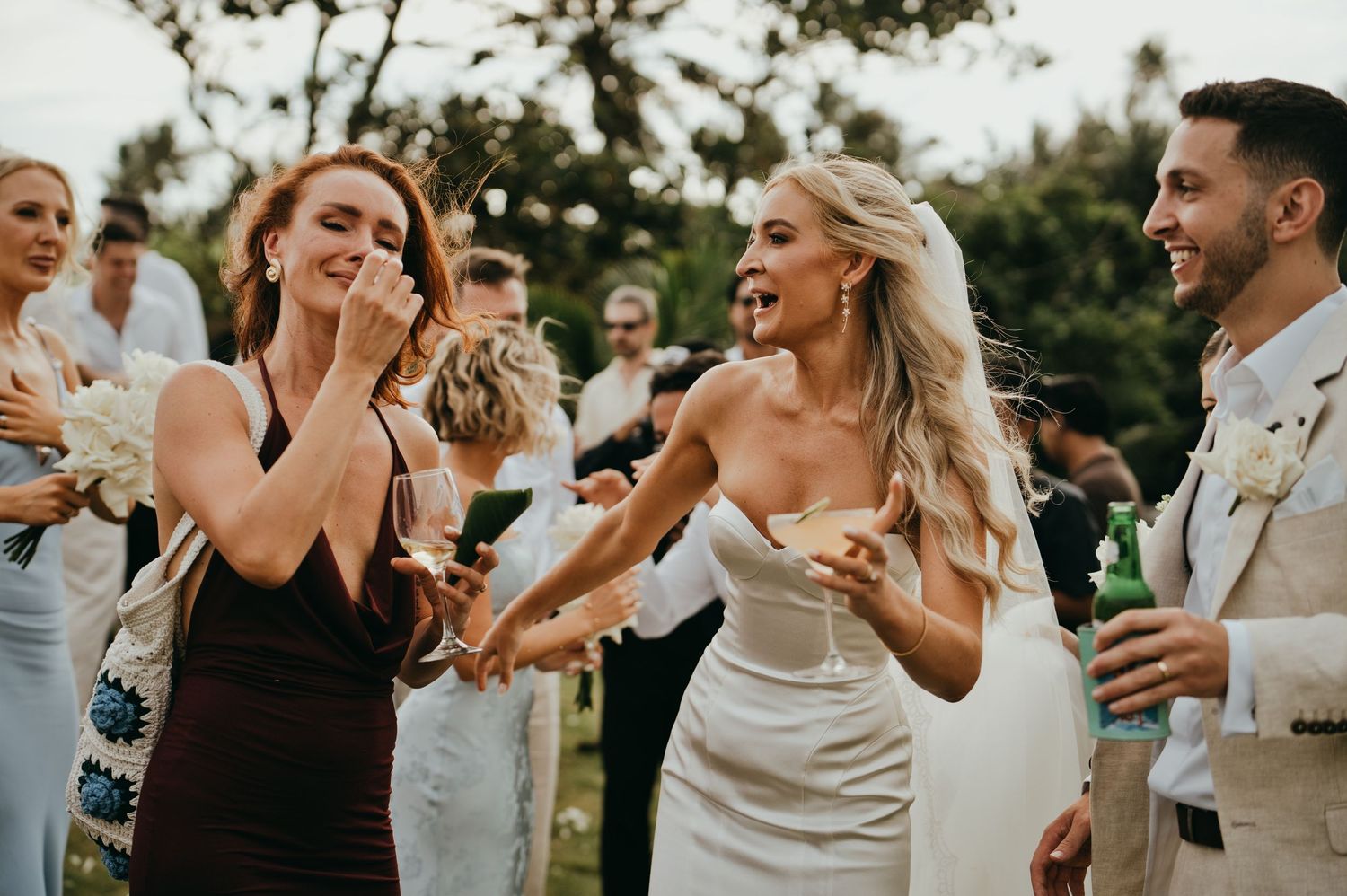 Laughing bride in white gown celebrates with bridesmaid in black dress during outdoor wedding cocktail hour.