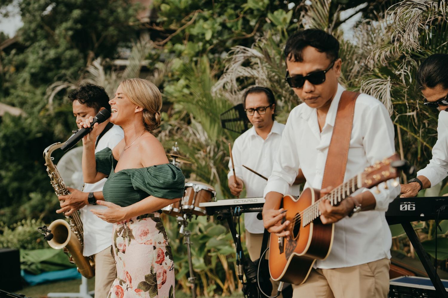 Live wedding band performing outdoors with saxophonist, guitarist, and vocalist in tropical setting.