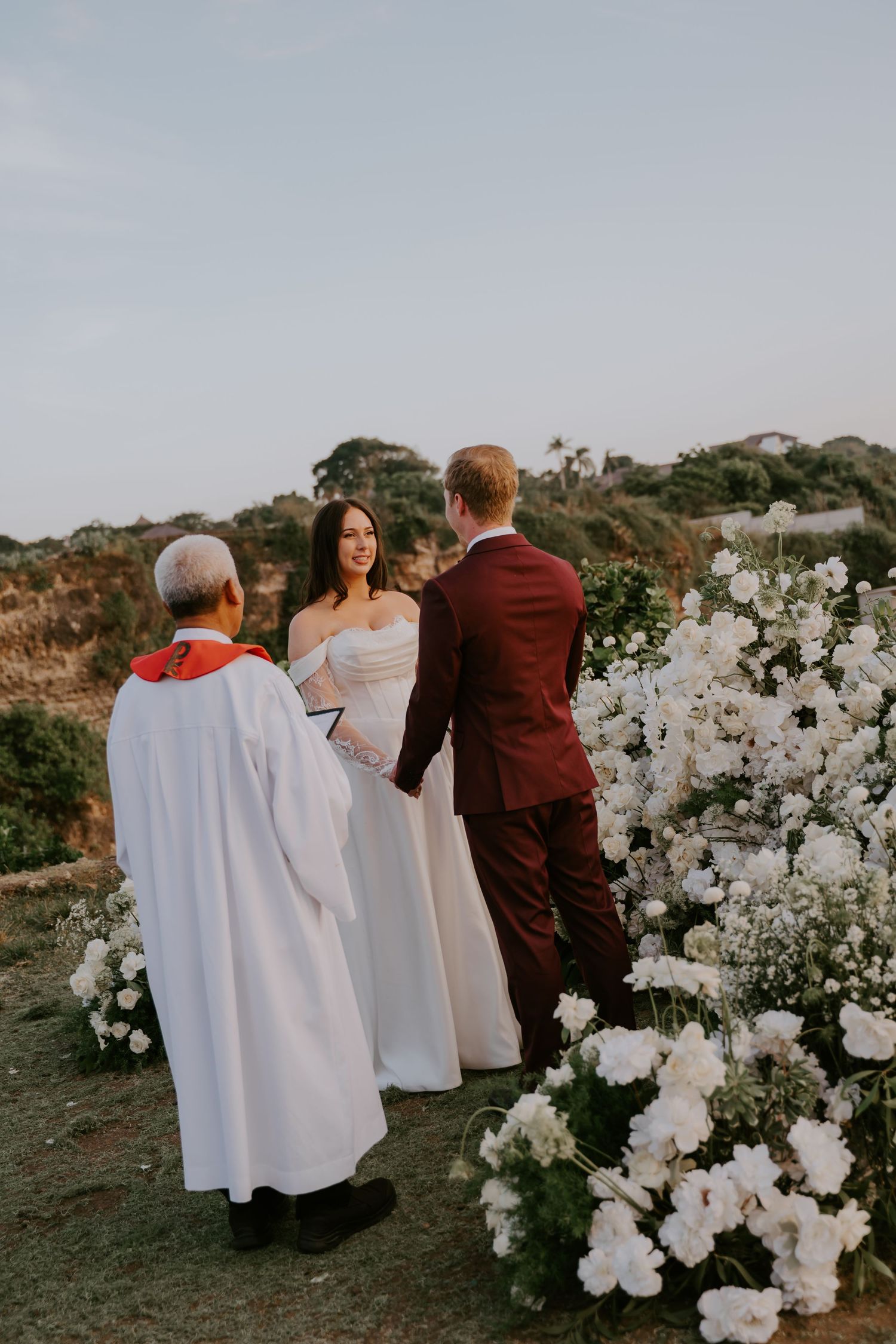 Couple exchanges vows outdoors with officiant beside white floral arrangements overlooking the ocean.