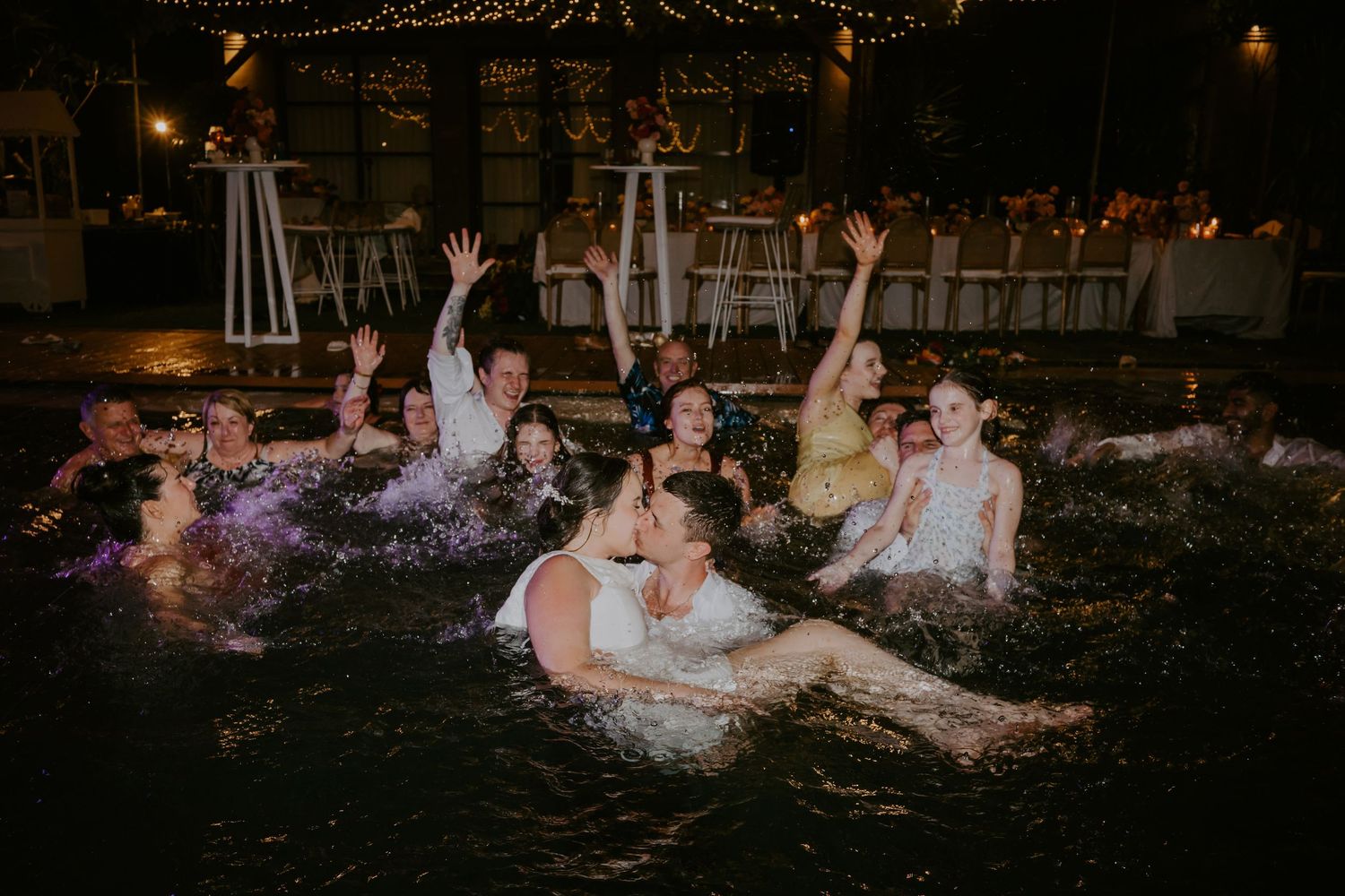 Bride, groom and wedding guests celebrating in illuminated pool during fun Bali wedding reception party.