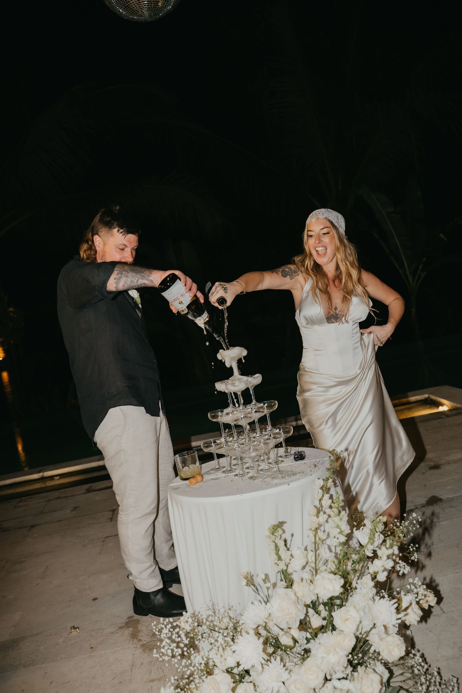 Bride and groom pour champagne tower at outdoor nighttime wedding celebration with white floral arrangements.