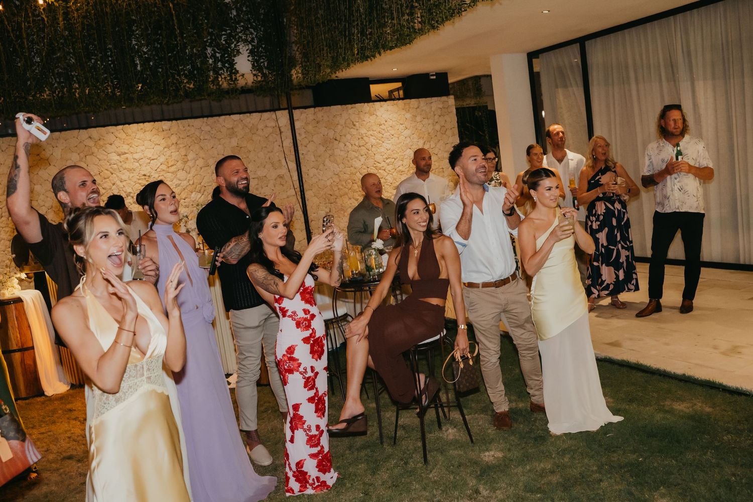 Wedding guests laugh and clap together during an outdoor evening reception celebration under string lights.