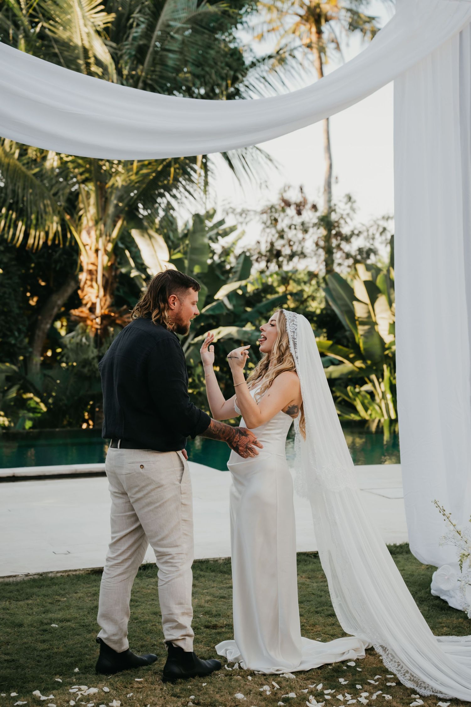 Bride and groom exchange vows under white draped arch surrounded by tropical palms at outdoor wedding ceremony.