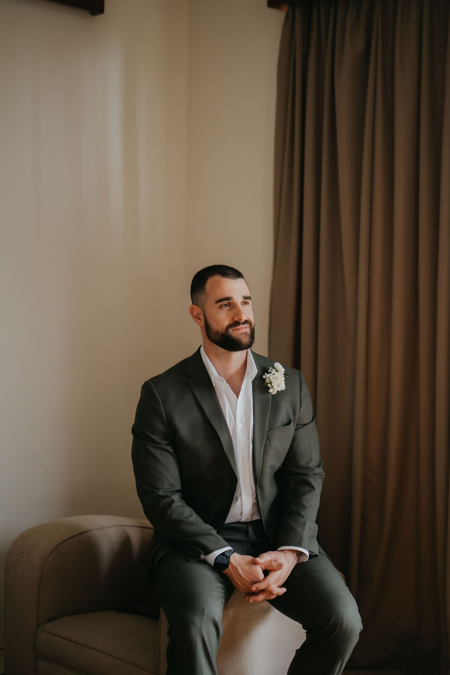 Groom in gray suit with boutonniere sits in armchair smiling before wedding ceremony.