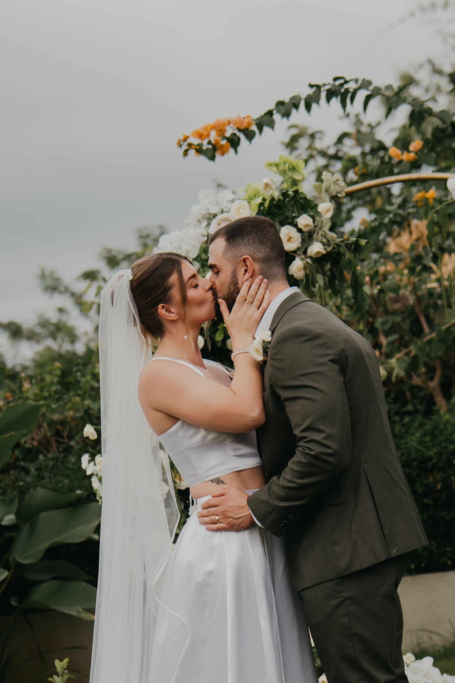Bride and groom share first kiss at outdoor wedding altar decorated with white flowers and tropical greenery.