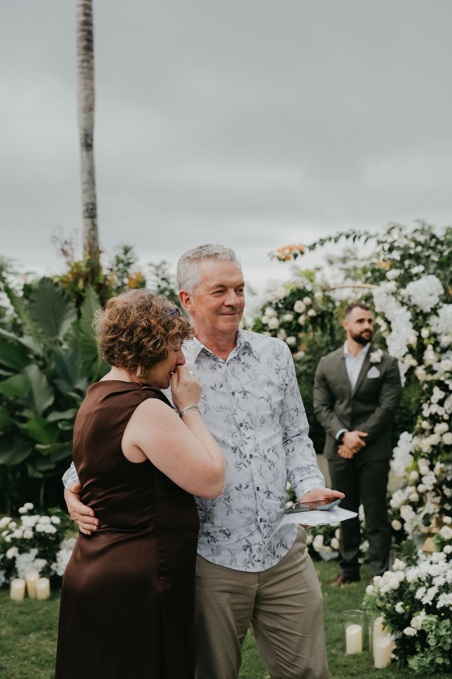 Emotional outdoor wedding ceremony moment with couple embracing near a floral arch and smiling groom.