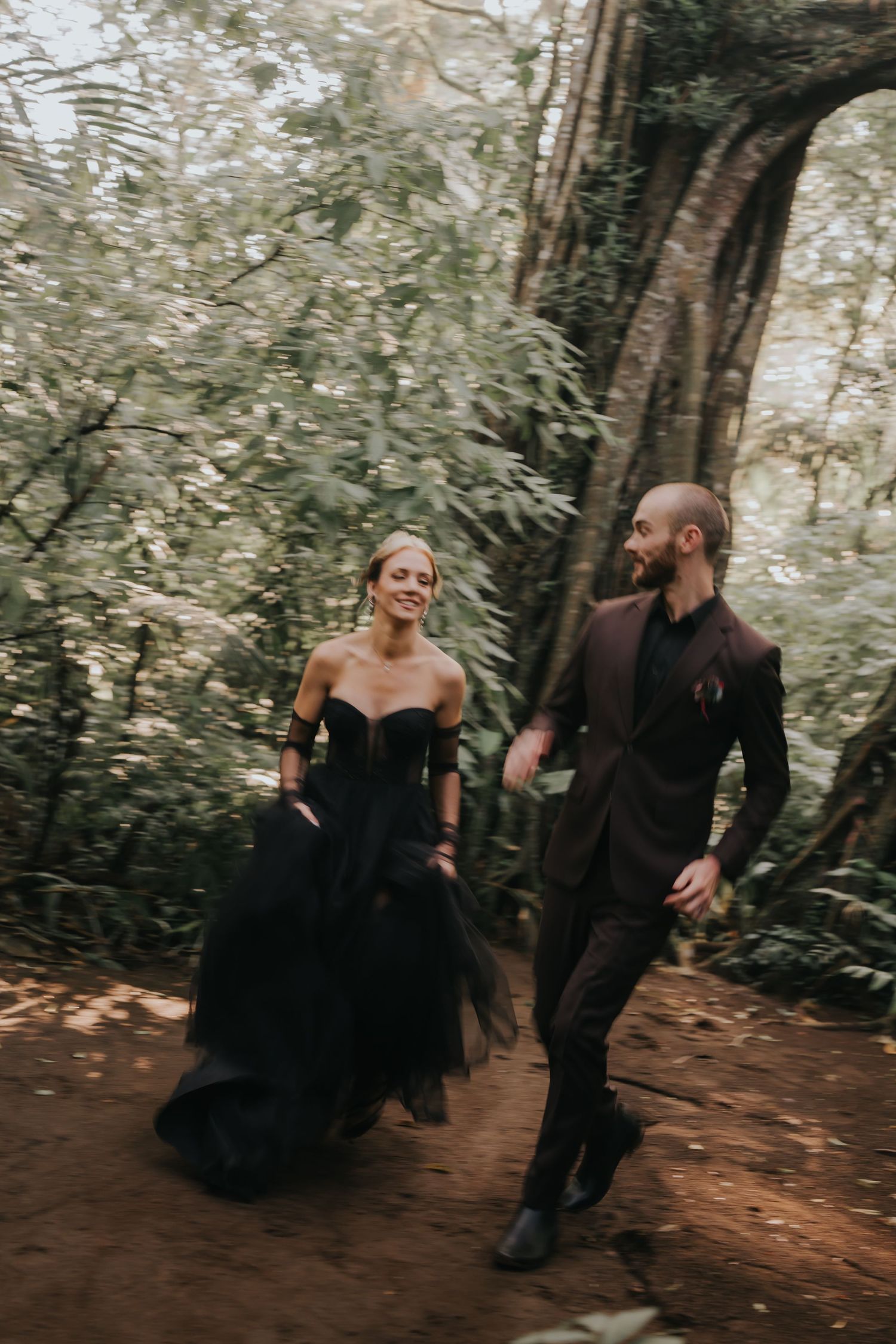 Couple in black formal wear walking and laughing together on a forest path.