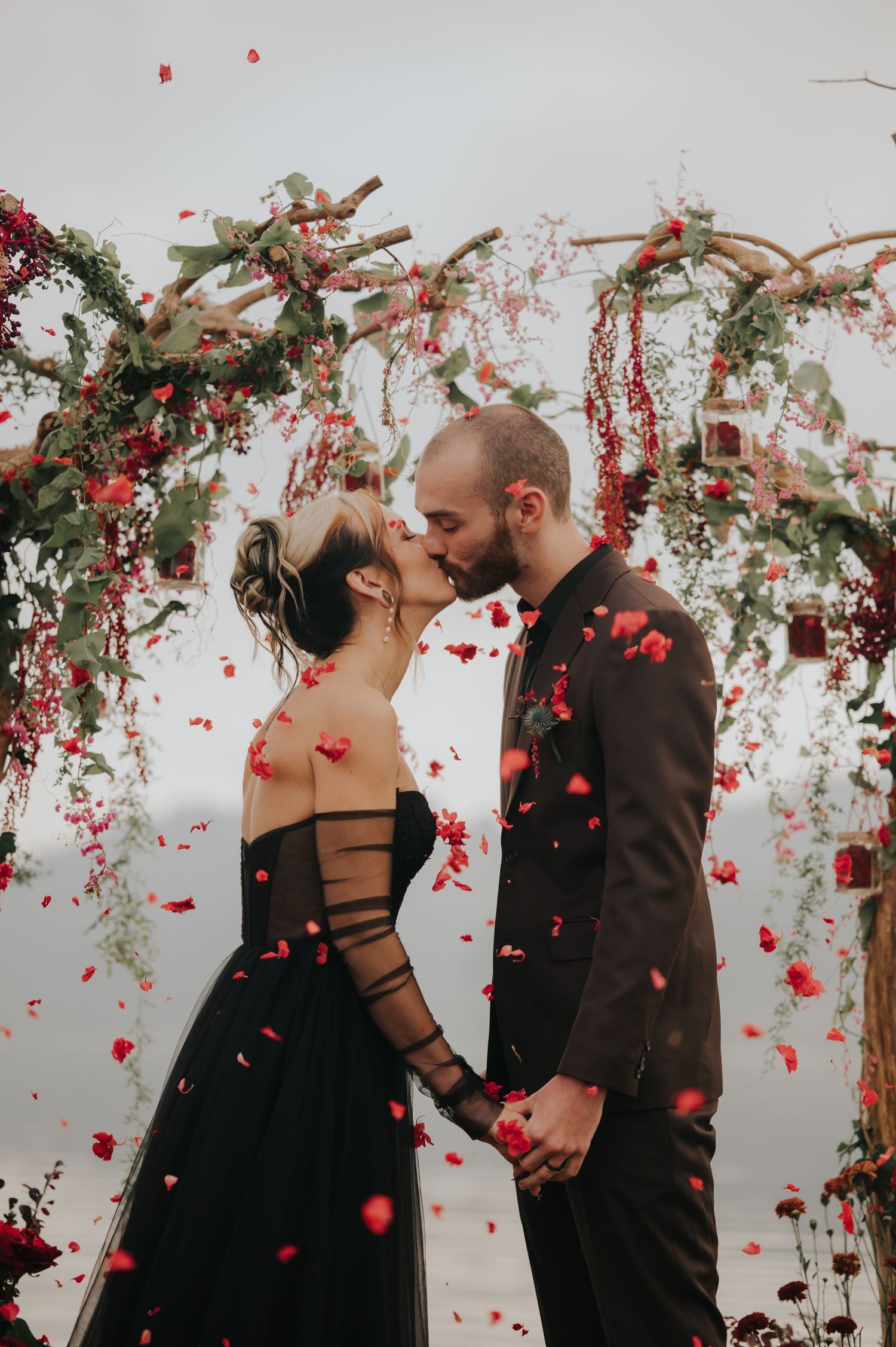 A romantic couple in black attire shares intimate moments under a floral arch as red rose petals fall around them.