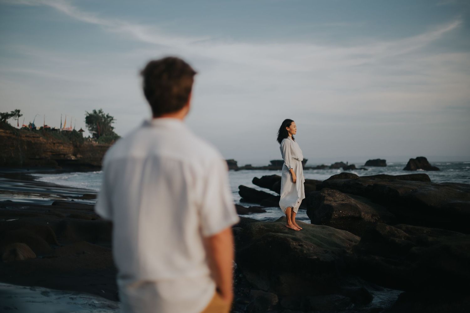 A couple walks along rocky coastal cliffs at sunset as waves crash against the shore in the background.