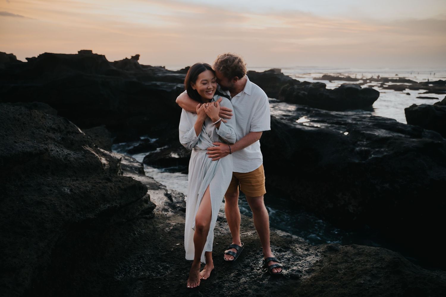 A couple embraces on rocky coastal cliffs at sunset, creating a romantic silhouette against the ocean backdrop.