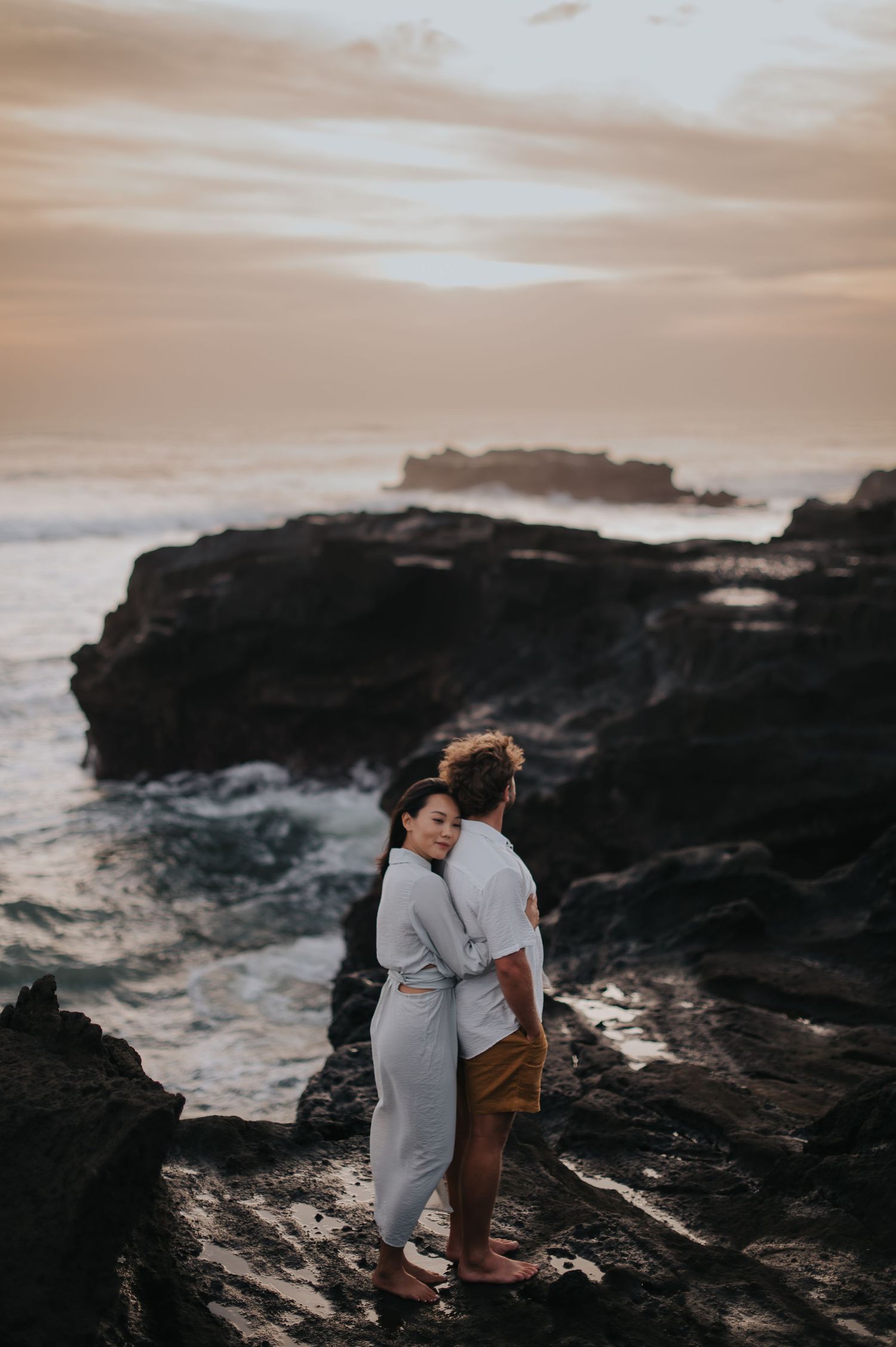 A couple shares an intimate moment on dark rocky cliffs overlooking the ocean at sunset.
