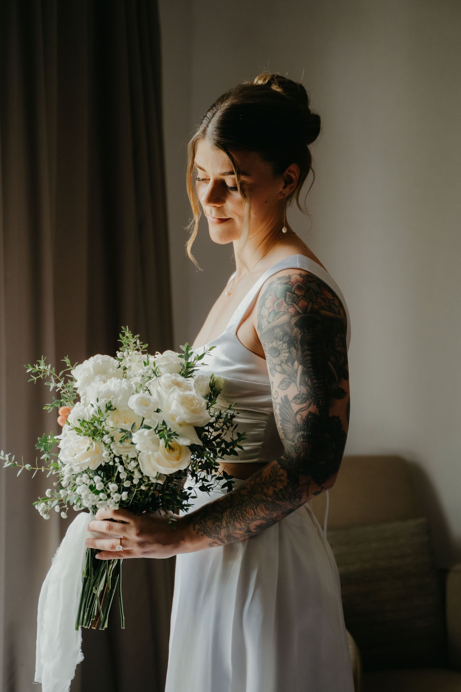 Tattooed bride in white gown holding elegant white floral bouquet by sunlit window.