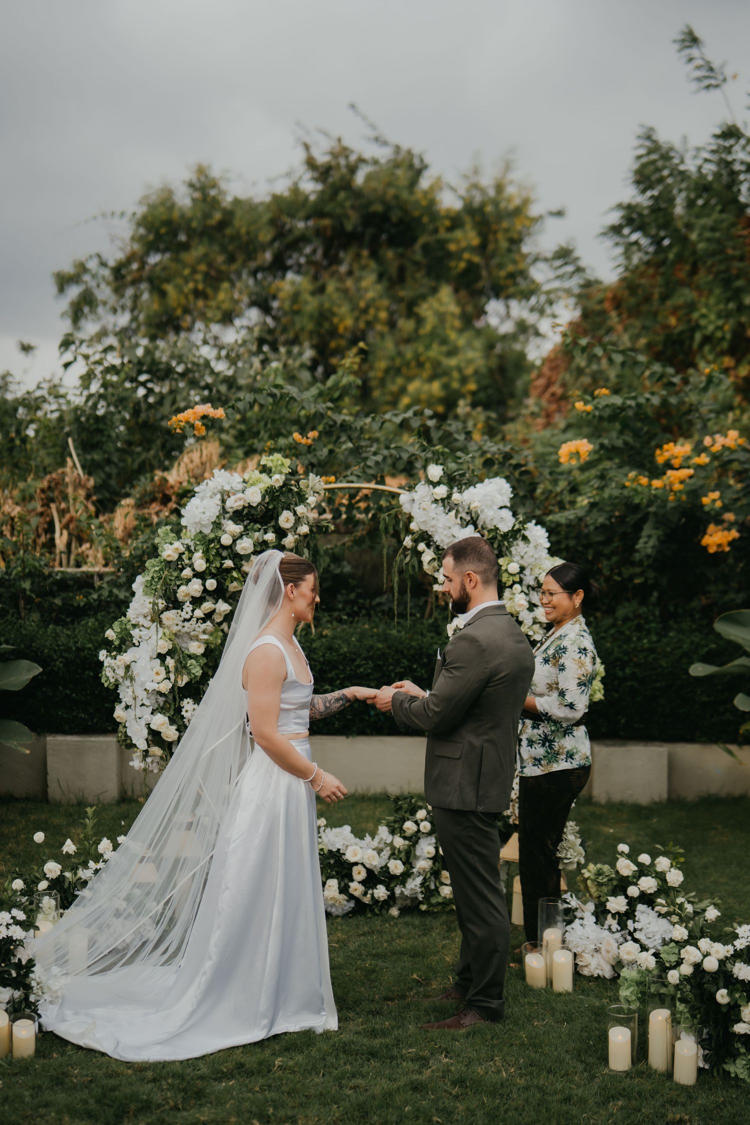 Couple exchanging vows under white floral arch with candles and officiant in garden.