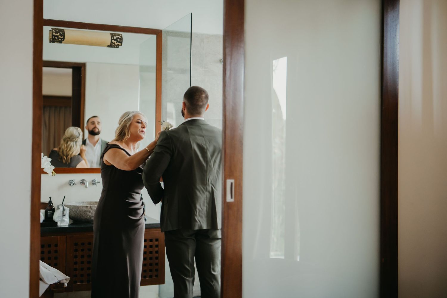 Couple reflected in bathroom mirror as woman helps man with his suit jacket before wedding.