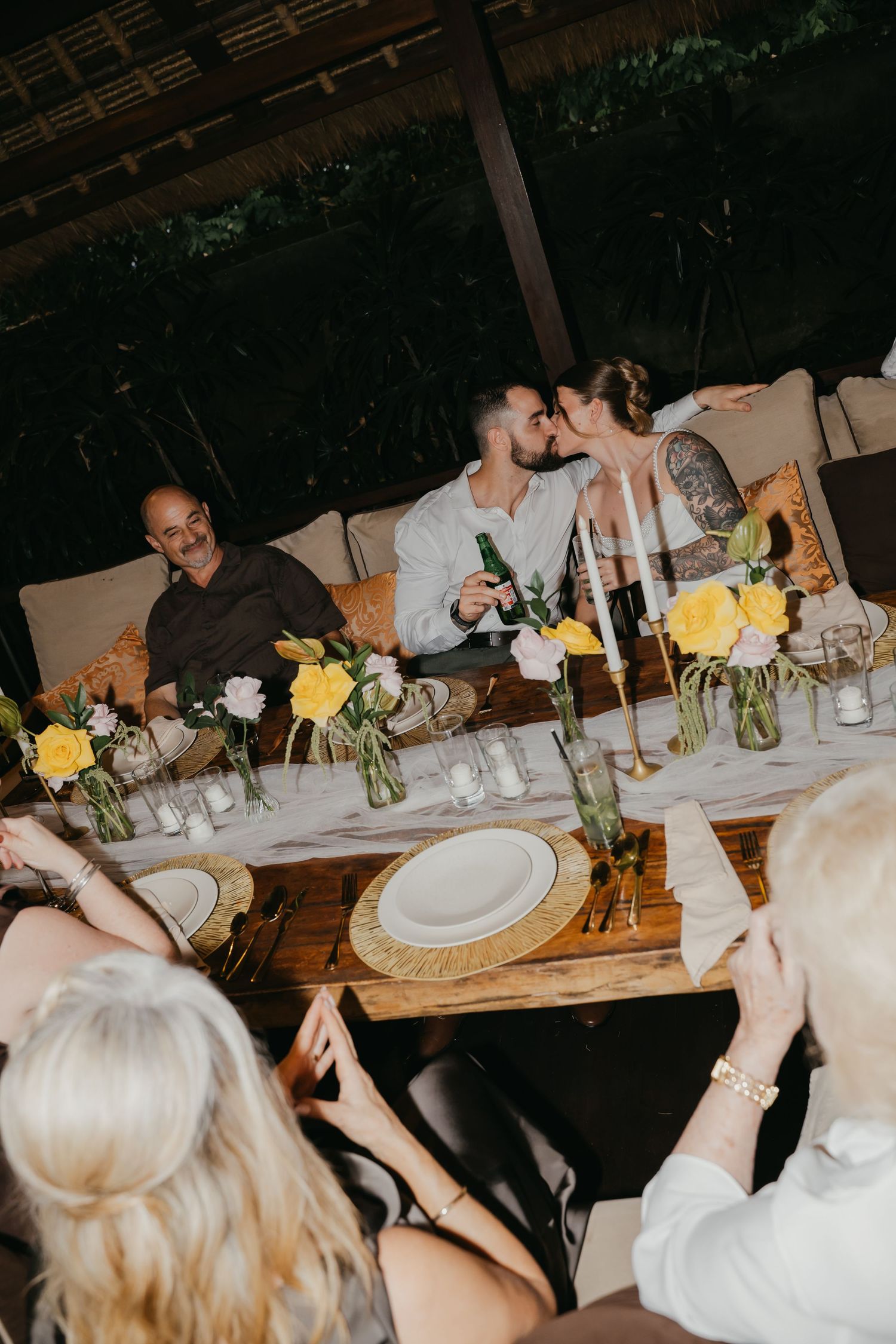 Newlyweds share a kiss at elegant outdoor wedding reception dinner table adorned with yellow floral arrangements.