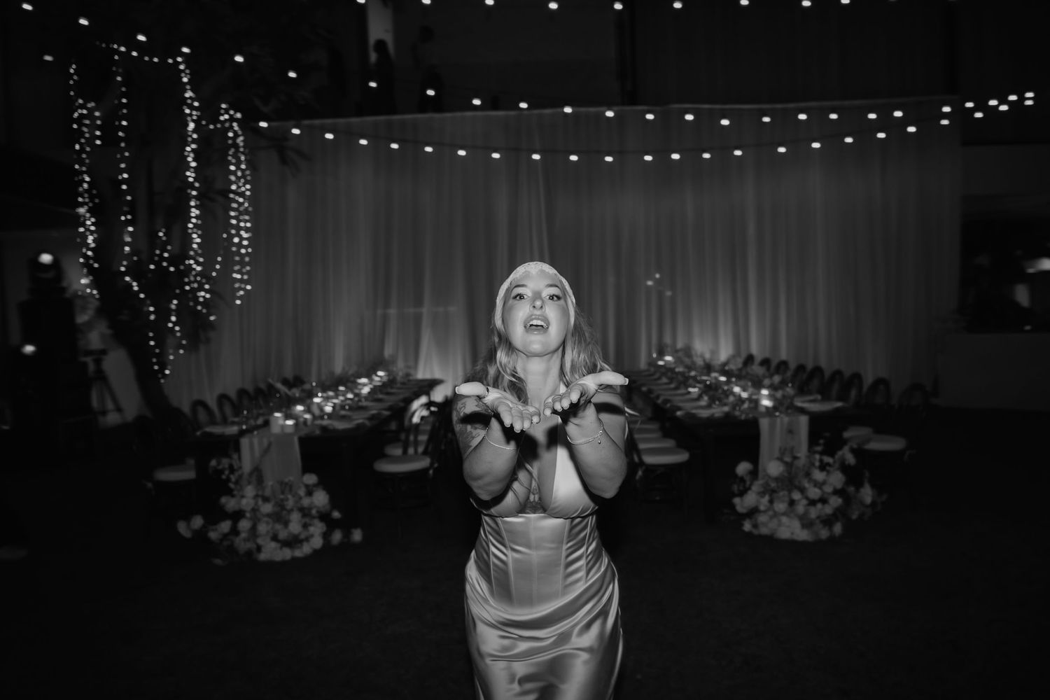 Woman in elegant dress blows a kiss in black and white photo at decorated wedding reception venue.