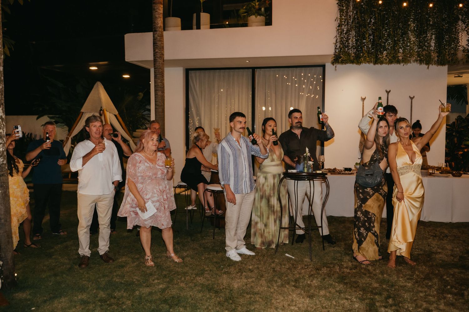 Wedding guests raising glasses for a toast outdoors at night during reception celebration.