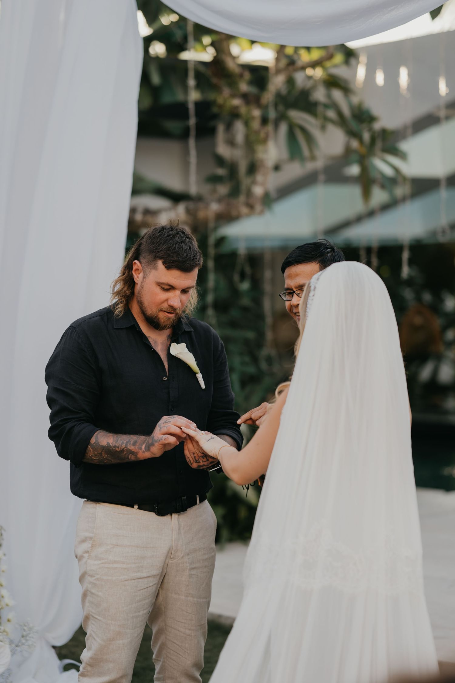 Groom placing wedding ring on brides finger during outdoor ceremony under white draped altar.