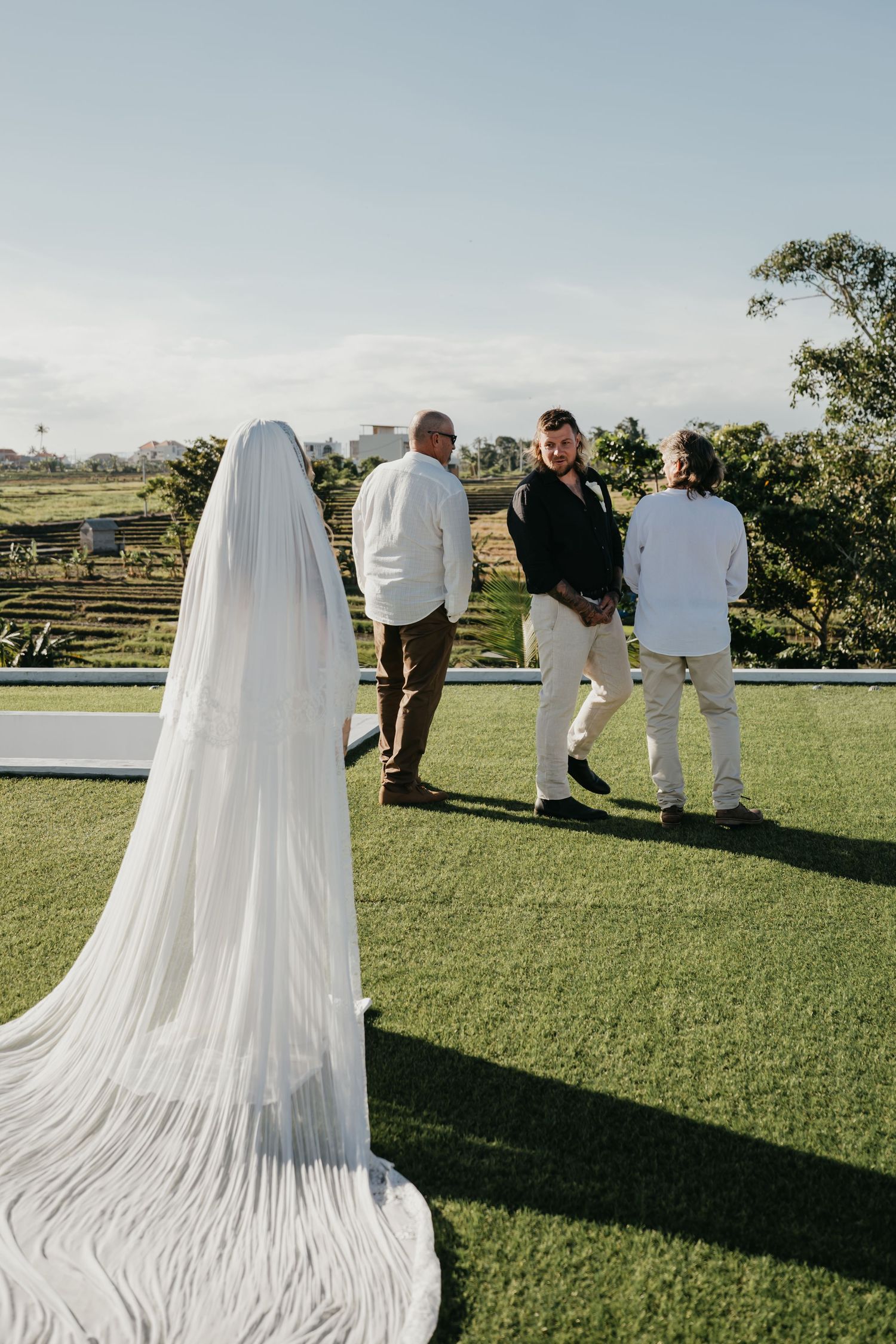 Bride in white gown and veil stands on green lawn as groom greets guests at outdoor wedding ceremony.