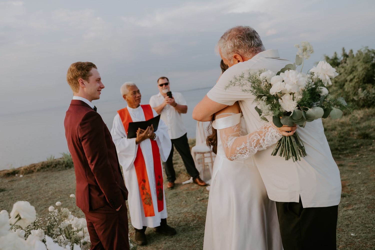 Outdoor wedding ceremony by the sea with bride hugging guest, groom in burgundy suit, and officiant in red stole.