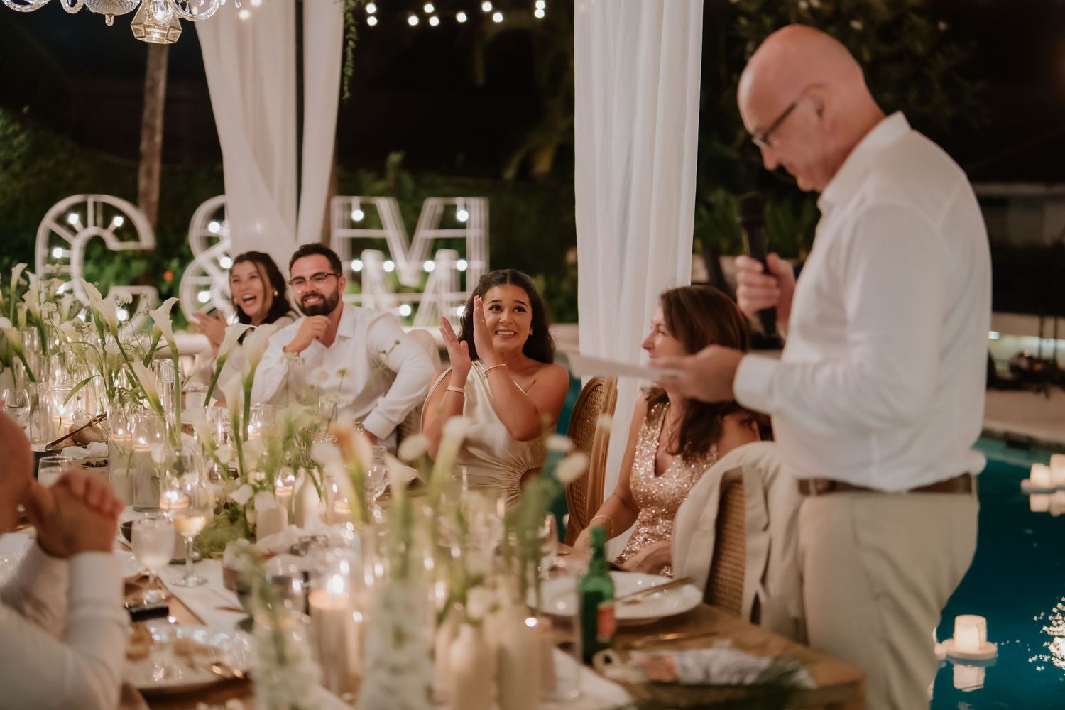 Wedding guests laughing during heartfelt speech at candlelit outdoor reception dinner.