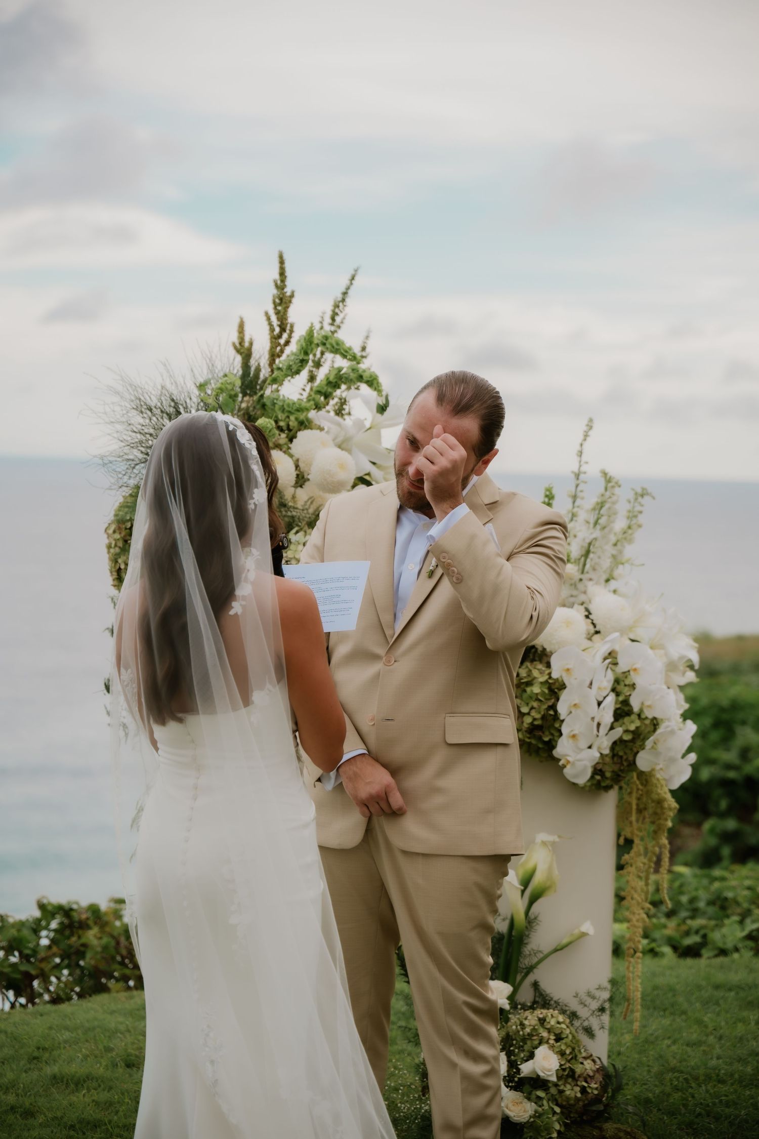 Emotional groom wiping tears as bride reads vows at outdoor wedding ceremony with white floral arch and ocean backdrop.