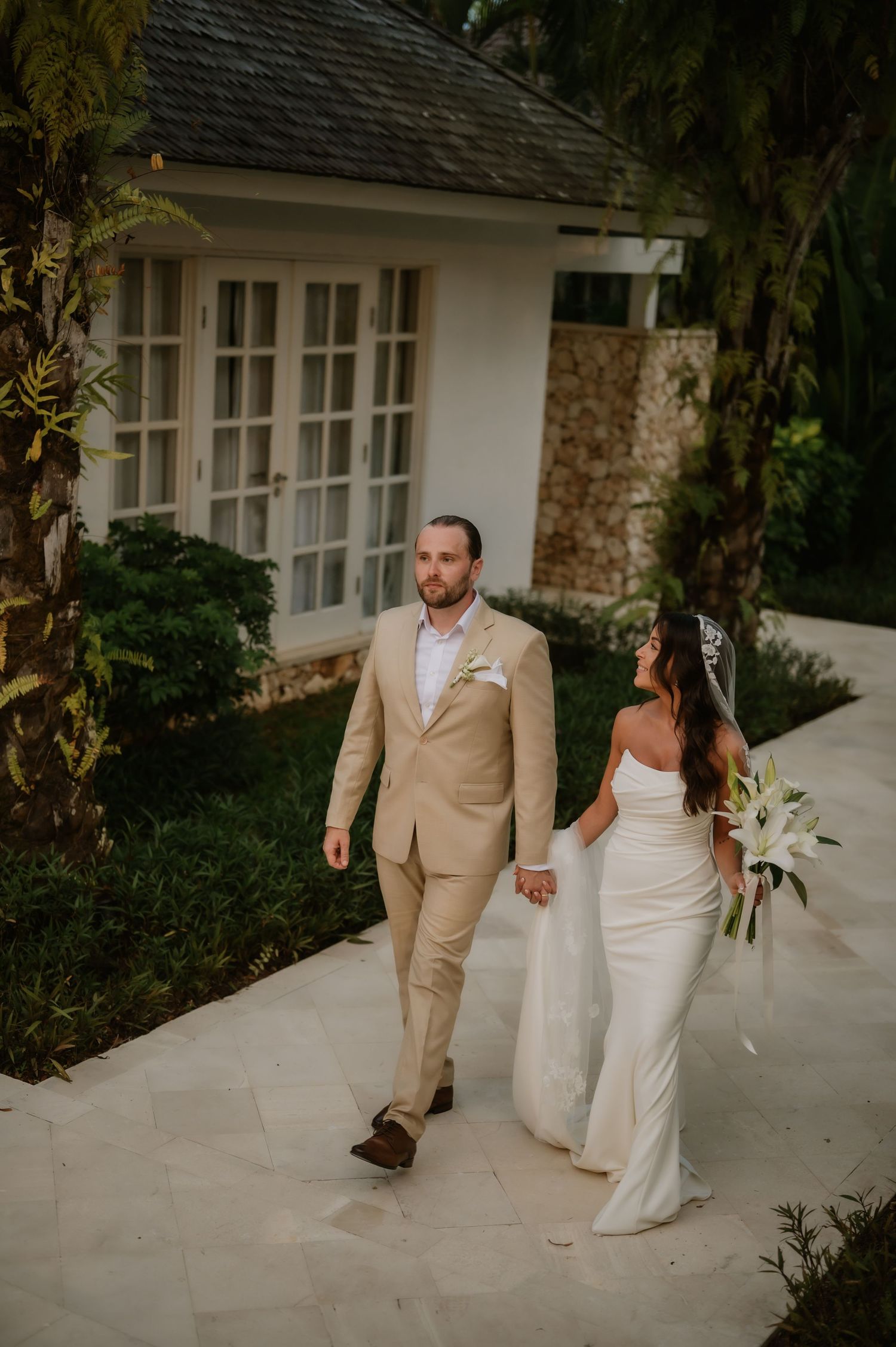 Bride in white gown and groom in tan suit walk hand-in-hand along a tropical villa pathway.