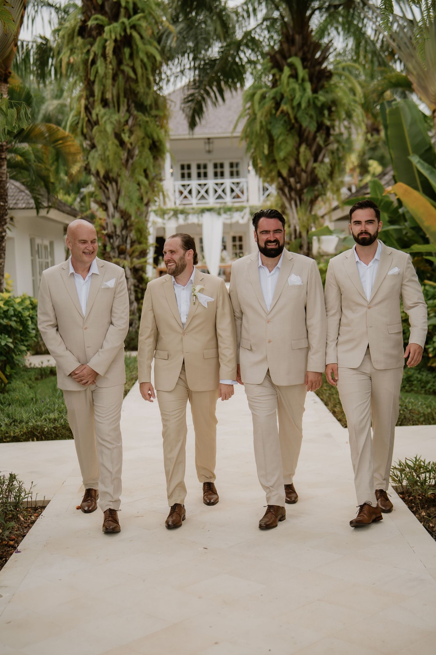 Four groomsmen in tan suits walking together on garden path with palm trees.