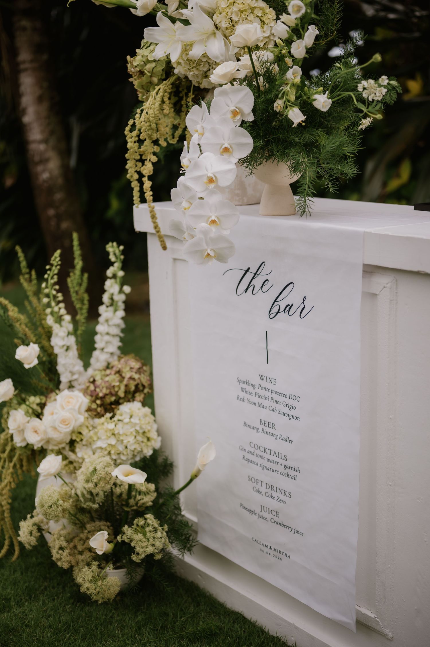 Elegant white wedding bar setup decorated with white orchids, hydrangeas, and a handwritten drink menu sign.