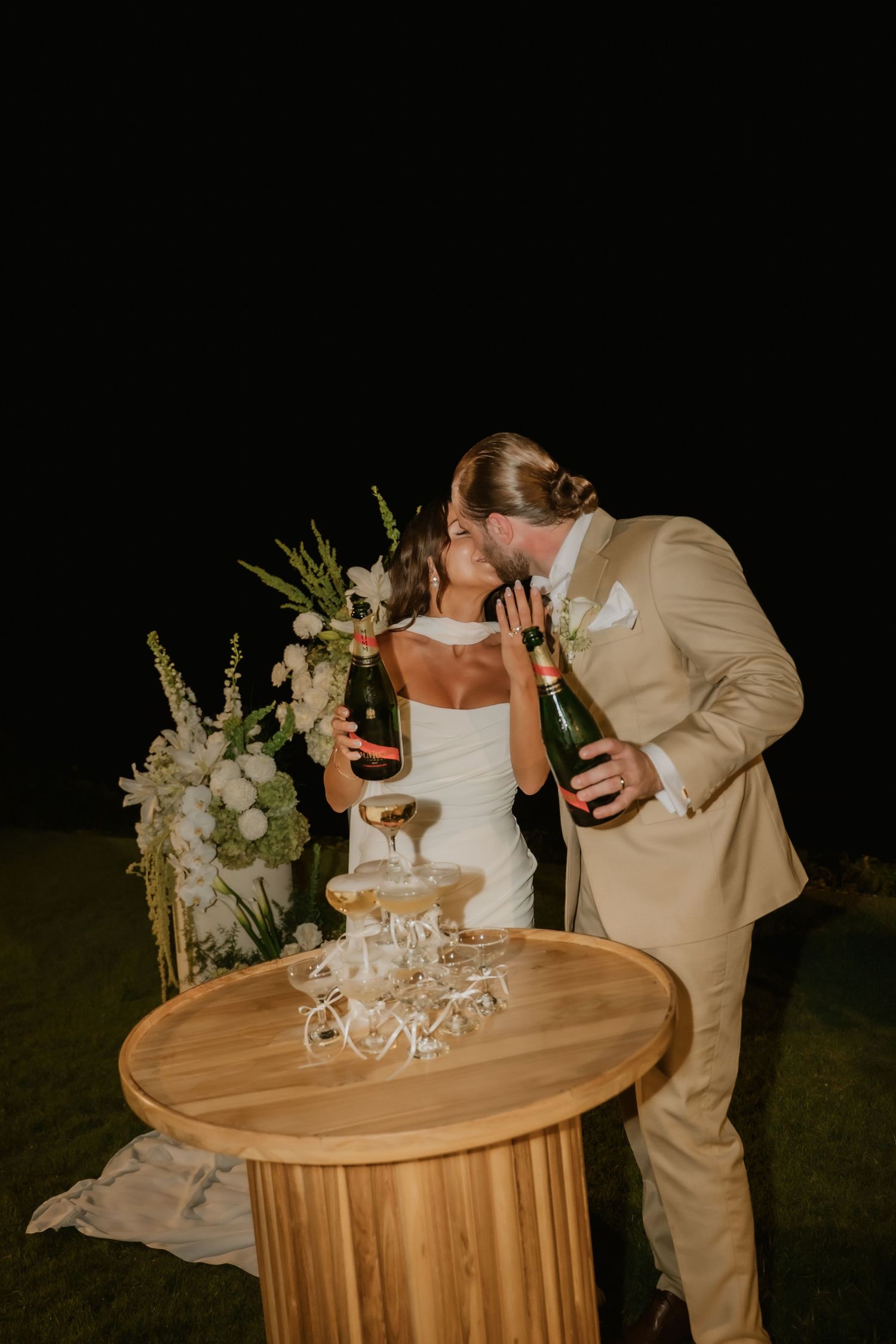 Bride and groom kissing while pouring champagne tower at outdoor wedding reception at night.