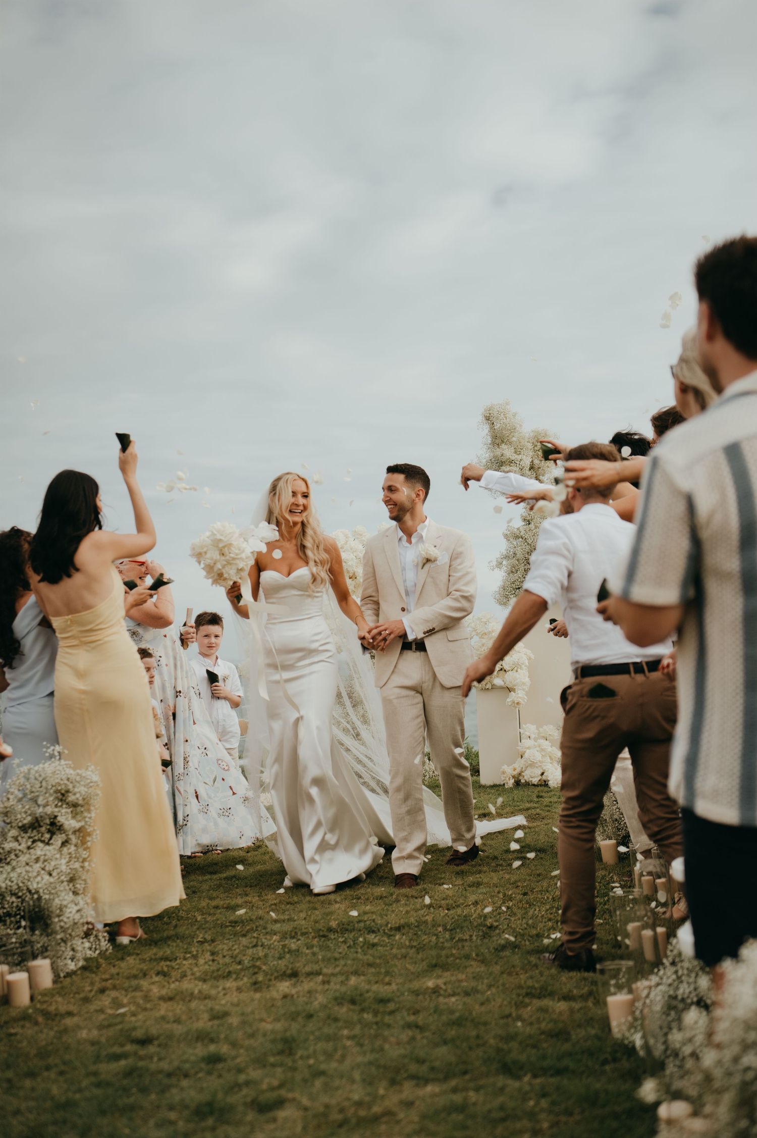 Joyful newlywed couple walking back down the aisle as guests celebrate with confetti at outdoor beach wedding.