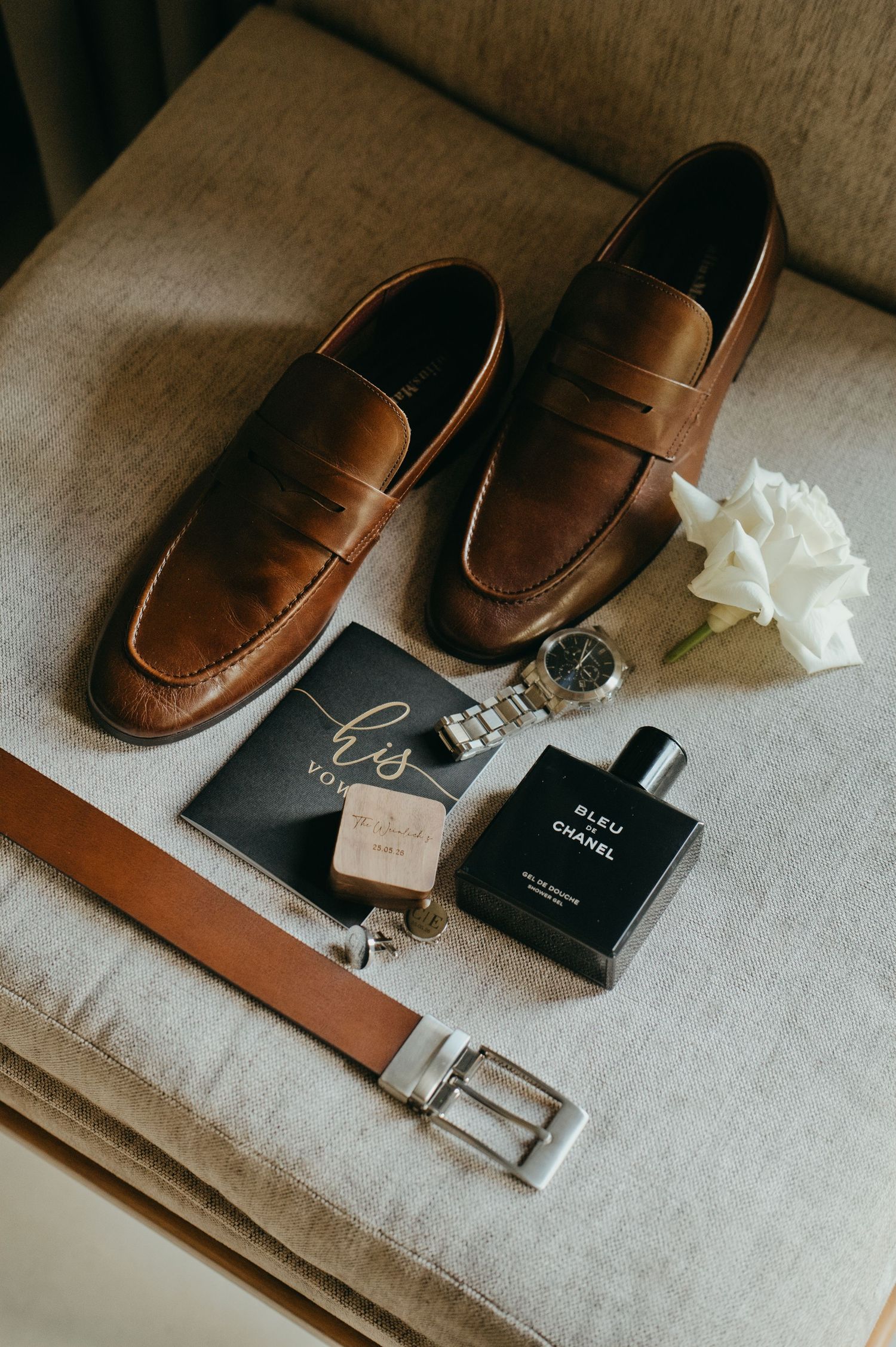 Brown leather loafers, Bleu de Chanel cologne, a watch, tan belt, and white roses styled as groom wedding accessories.